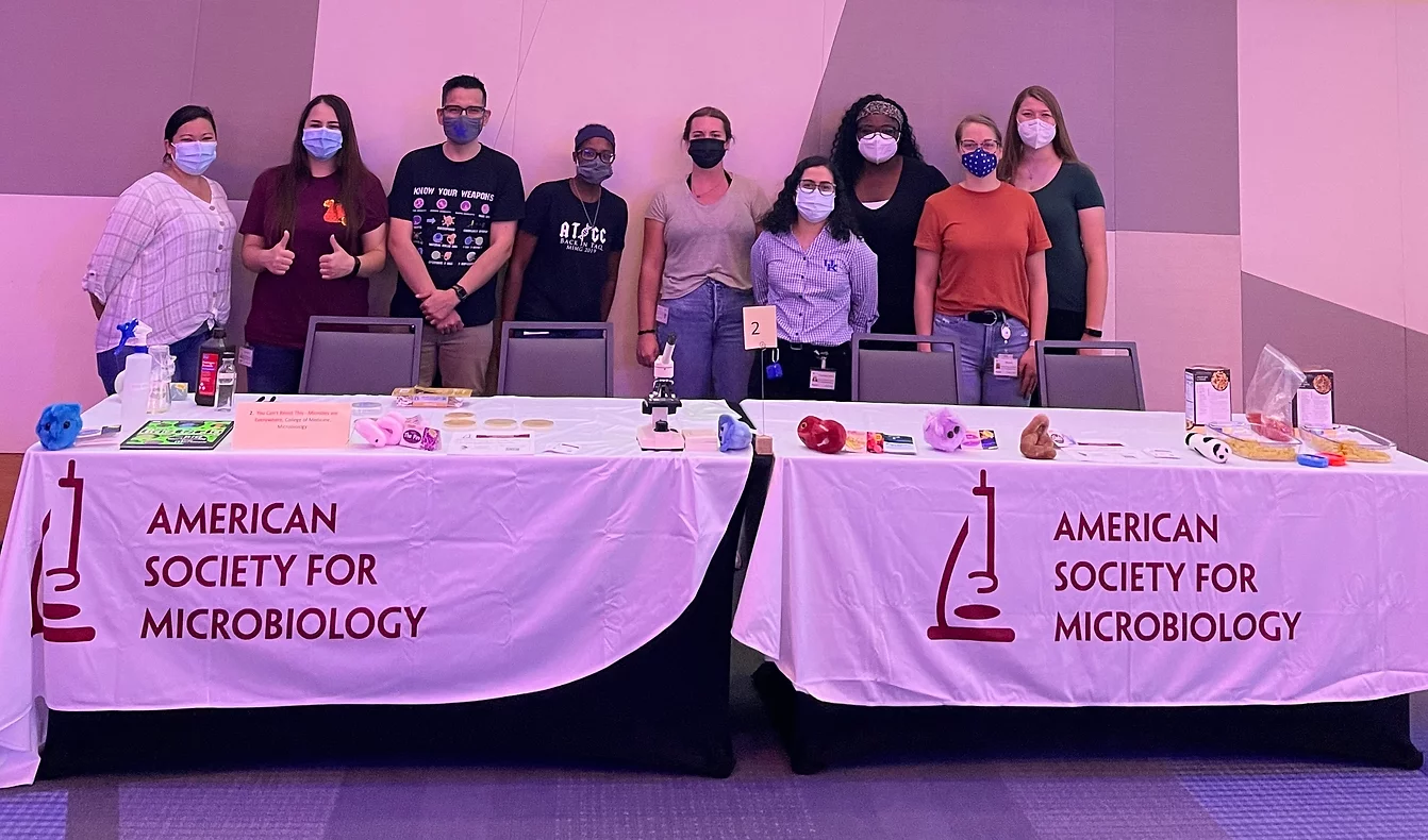 two display tables with a banners that say American Society for Microbiology with papers, petri dishes, microscope, pasta, and other stuff on the tables. Group of members behind the tables,
