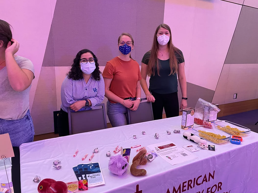 3 ASM student members standing behind a conference display table. Candy, brochures, stuffed animals, and pasta on the table.