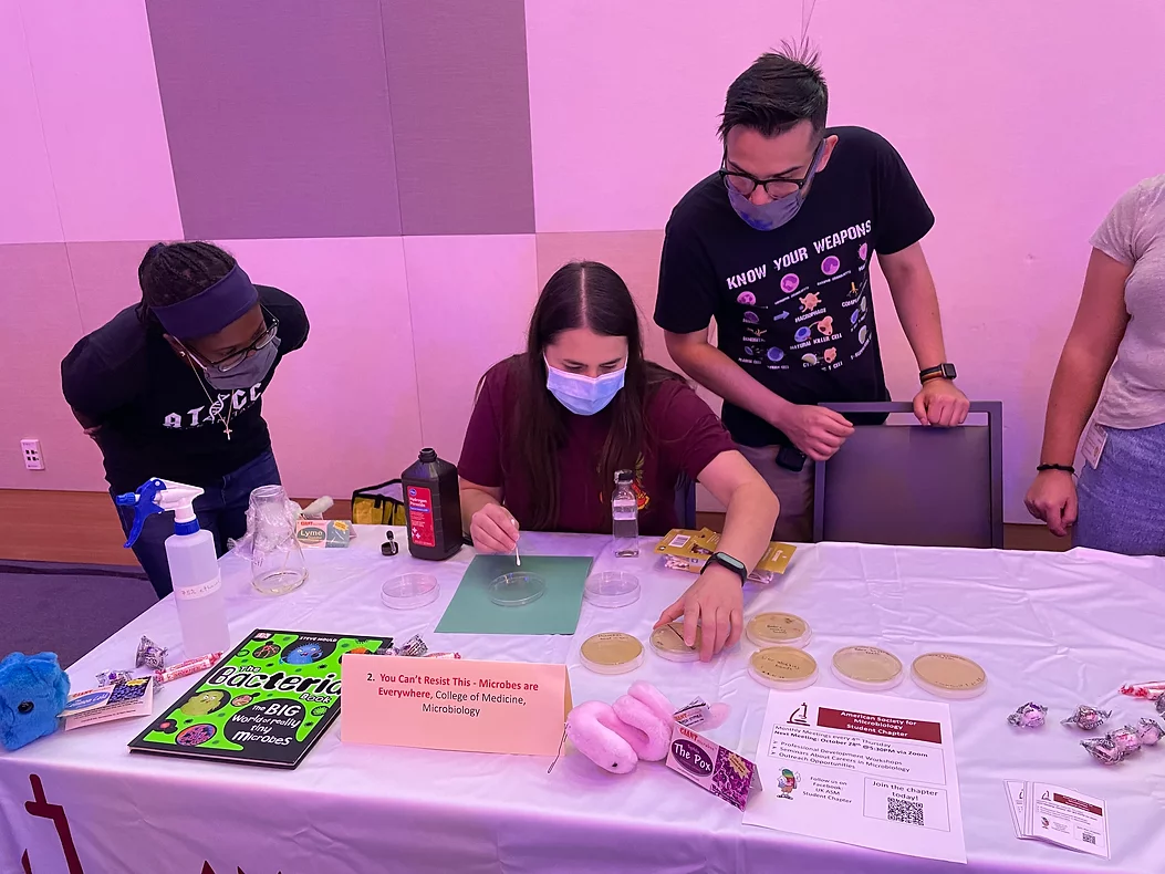 Demonstration table with petri dishes, The Bacteria Book, cleaning supplies, and 3 people looking down at the table