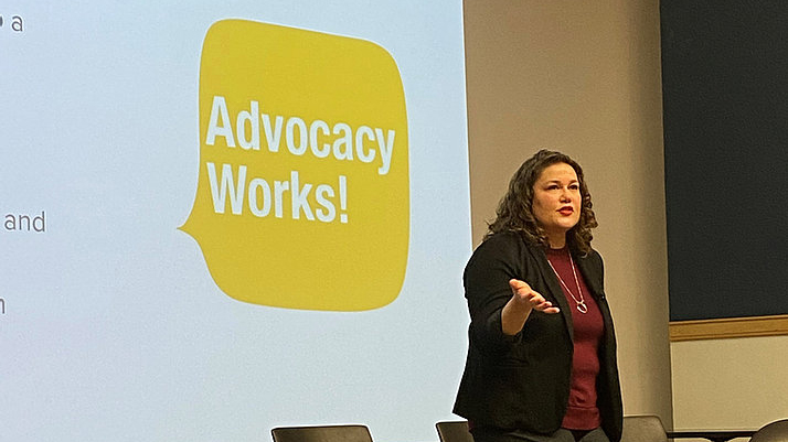 Katie McHugh, MD speaking on stage with an image in the background that says "Advocacy Works!"