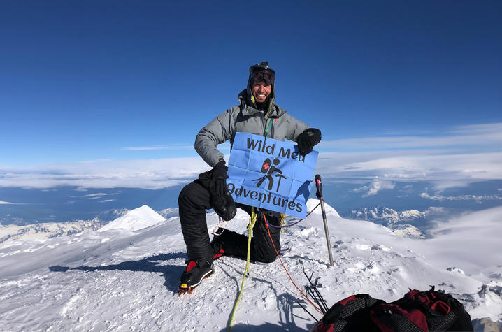 Ben Mattingly on Denali, the highest mountain peak in North America, holding a blue sign that says "Wild Med Adventures"
