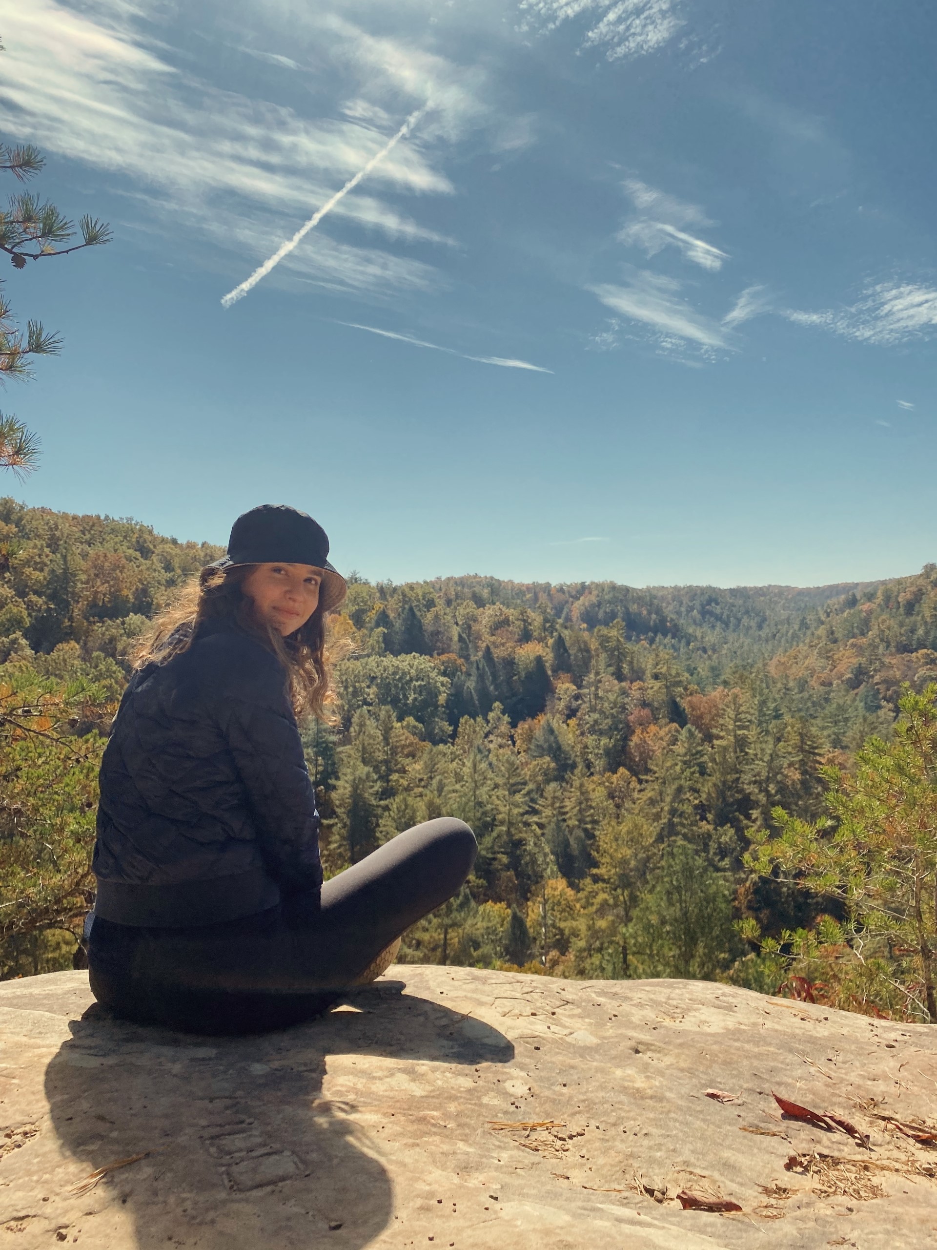 Diana Khair sitting at the top of a hiking lookout with the sky and tree landscape behind her. 
