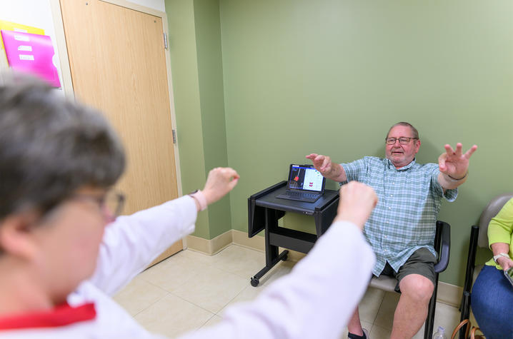 Julie Gurwell, a doctor, guiding Hoyt Ball, the patient, through an assessment where he mirrors her by raising his arms. Dr. Gurwell and Hoyt Ball sit in a green doctor's office as they screen him.