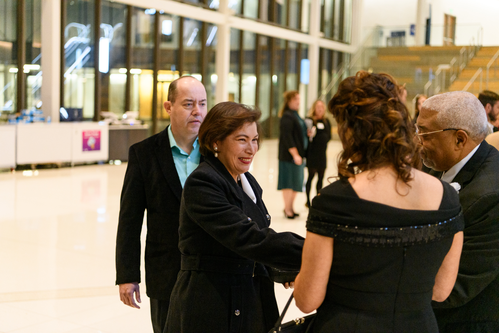 Two pairs approach each other in formal attire in a brightly lit foyer. A woman and man shake hands. 