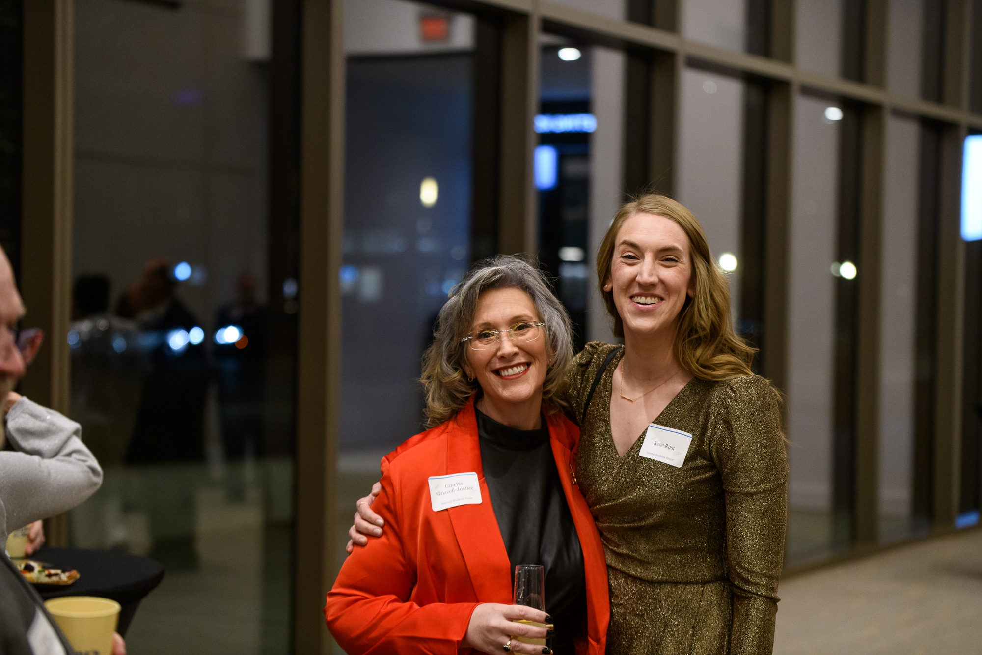 Two women smile for a photo in formal attire. The woman on the right wraps her arm around the other.