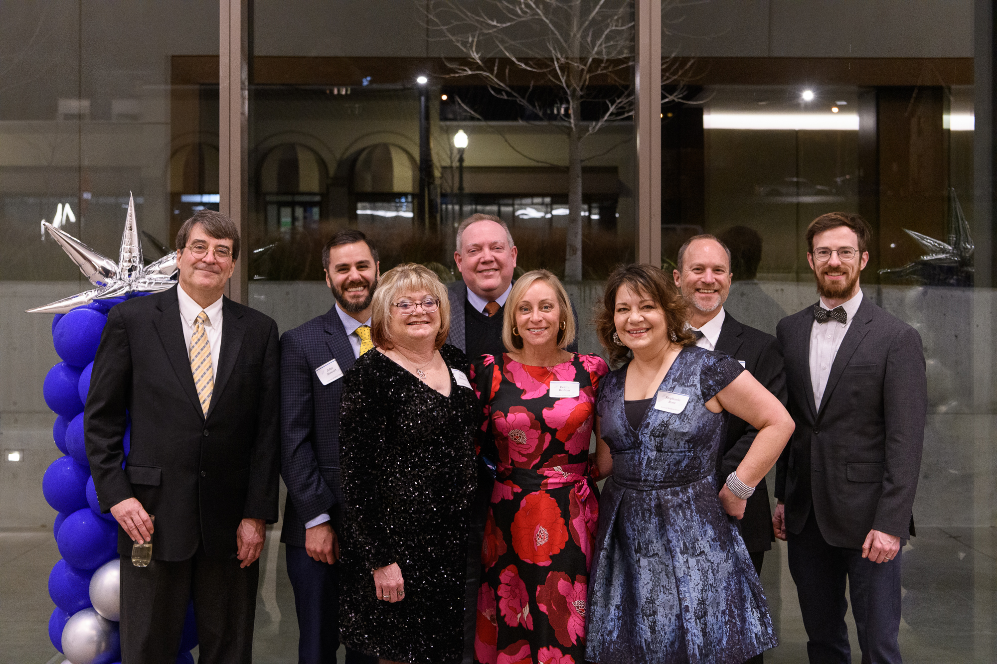 A group of people in formal attire smile for a photo. They stand in front of darkened windows