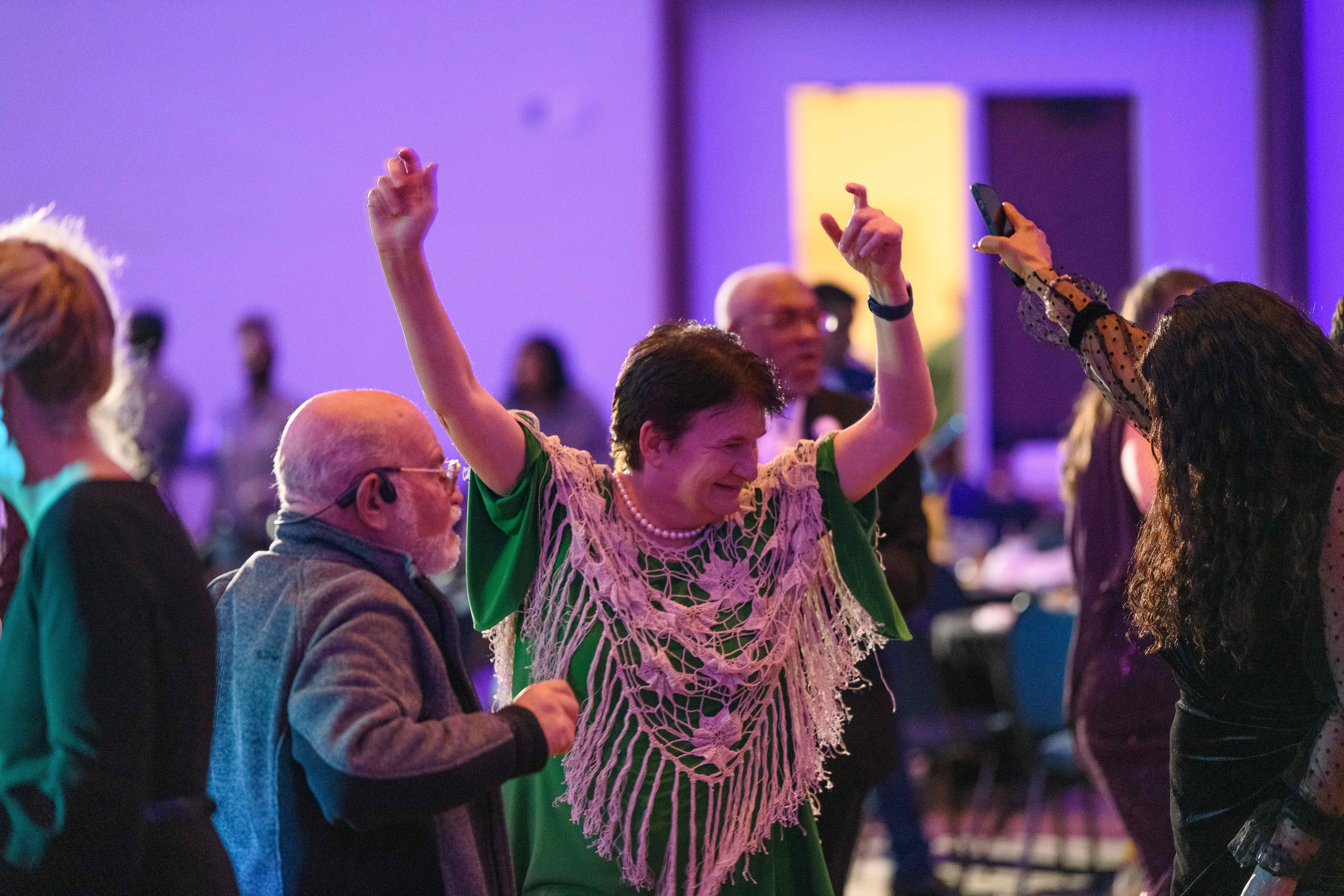 A woman in a green dress dances beneath colored lights, raising her arms up. Another woman cheers her on.