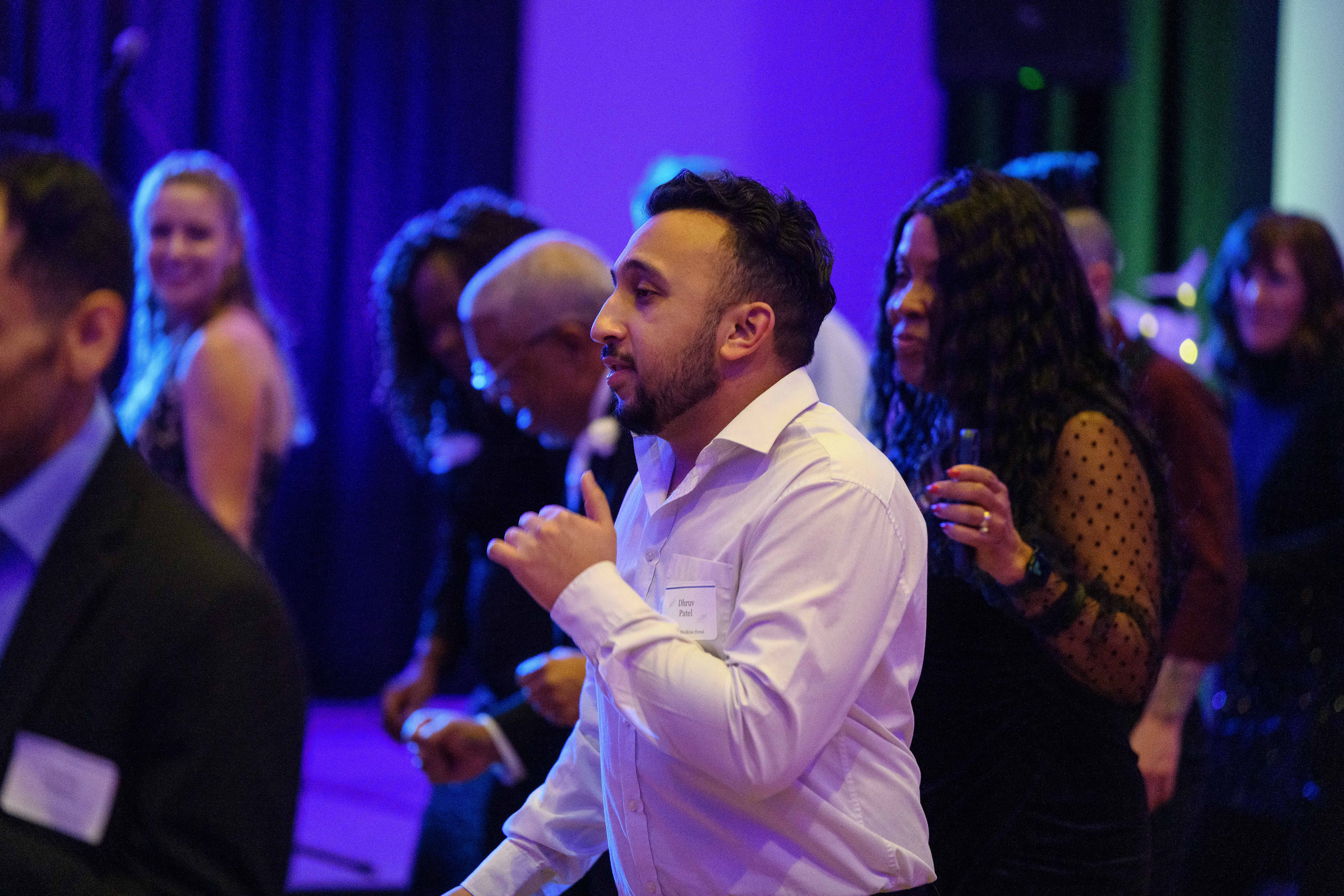 A group of people dance beneath colored lights. A man in a white shirt is the focal point.