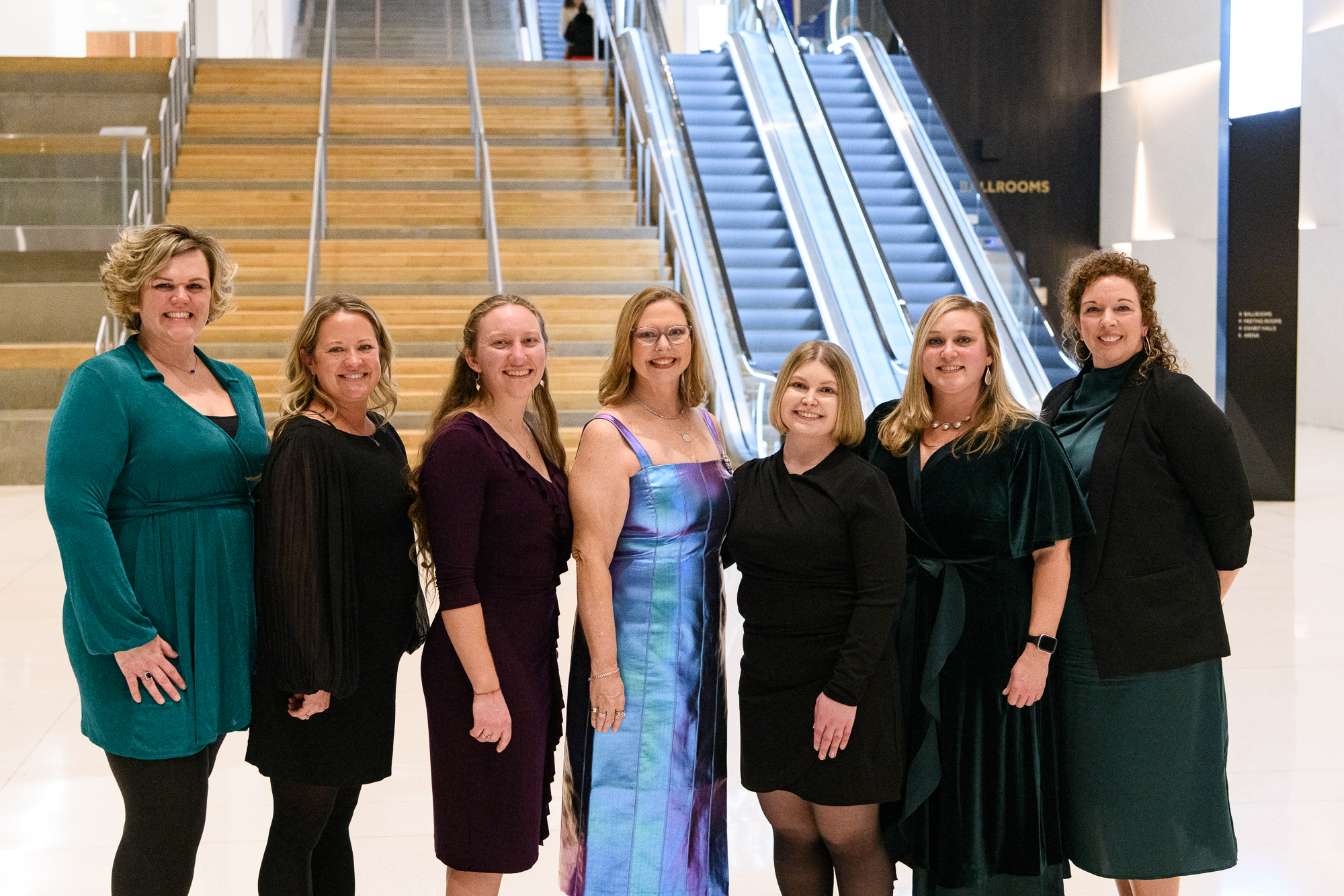 A group of women in formal attire stand in a line, uniformly posing and smiling at the camera.