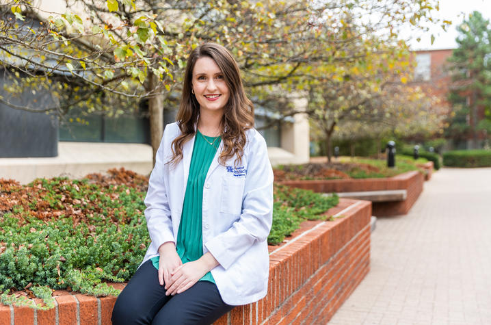 Susanna Goggans, wearing a white coat, sitting on the wall of a garden bed on UK's campus.