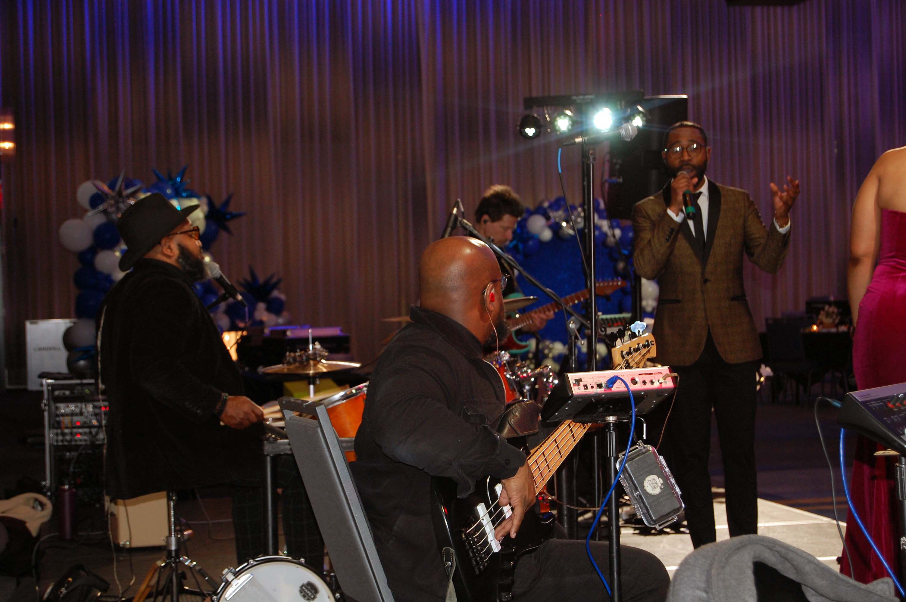 A band upon a stage perform amidst sound equipment. There are two guitarists, a drummer, and a singer