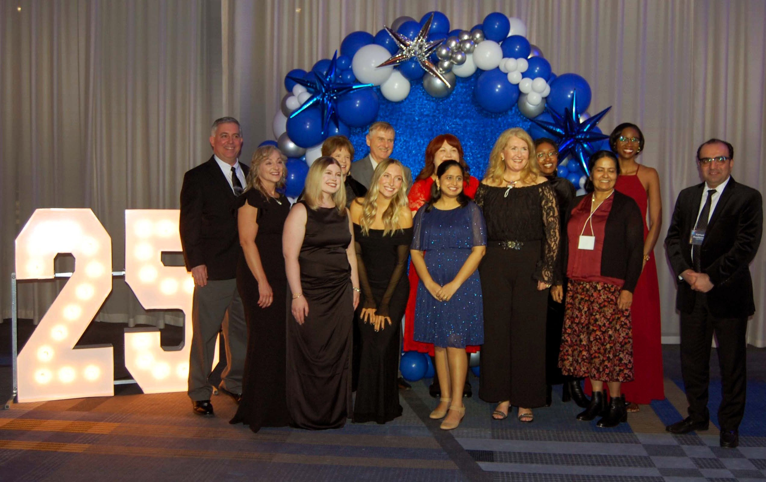A large group of people in formal attire pose for a photo. They stand in front of a balloon arch and big, glowing numbers that say 25