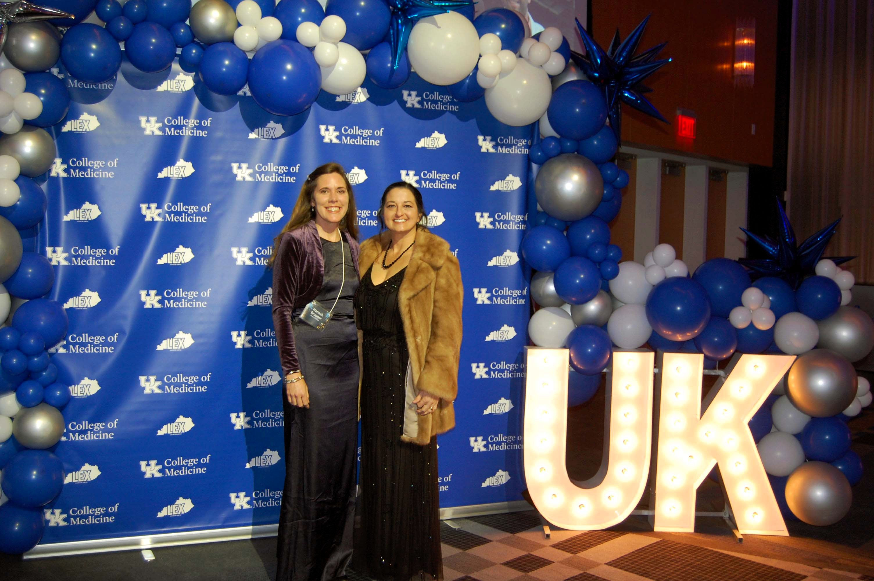 Two women in formal attire smile in front of a balloon arch. They stand next to big, glowing letters that say UK