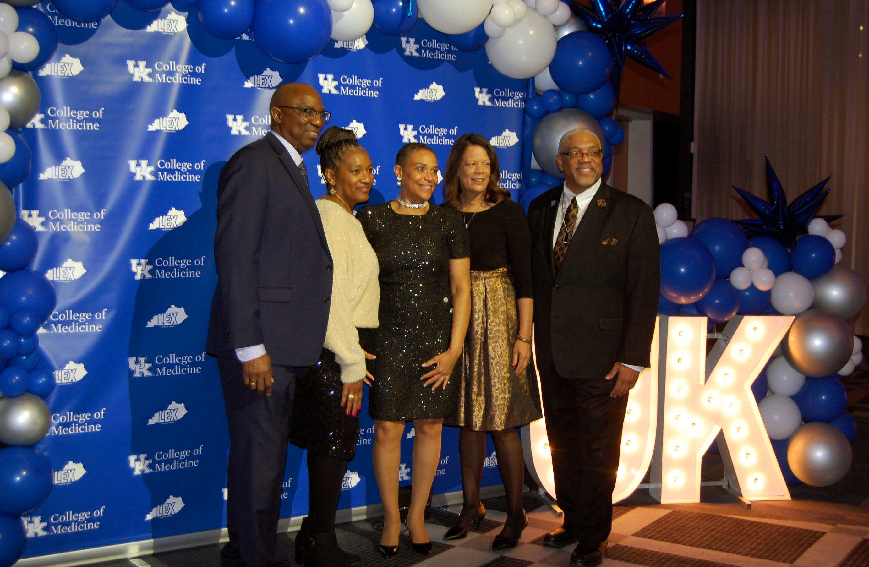 A group of five people in formal attire smile for a photo. They stand next to a balloon arch and big, glowing letters that say UK