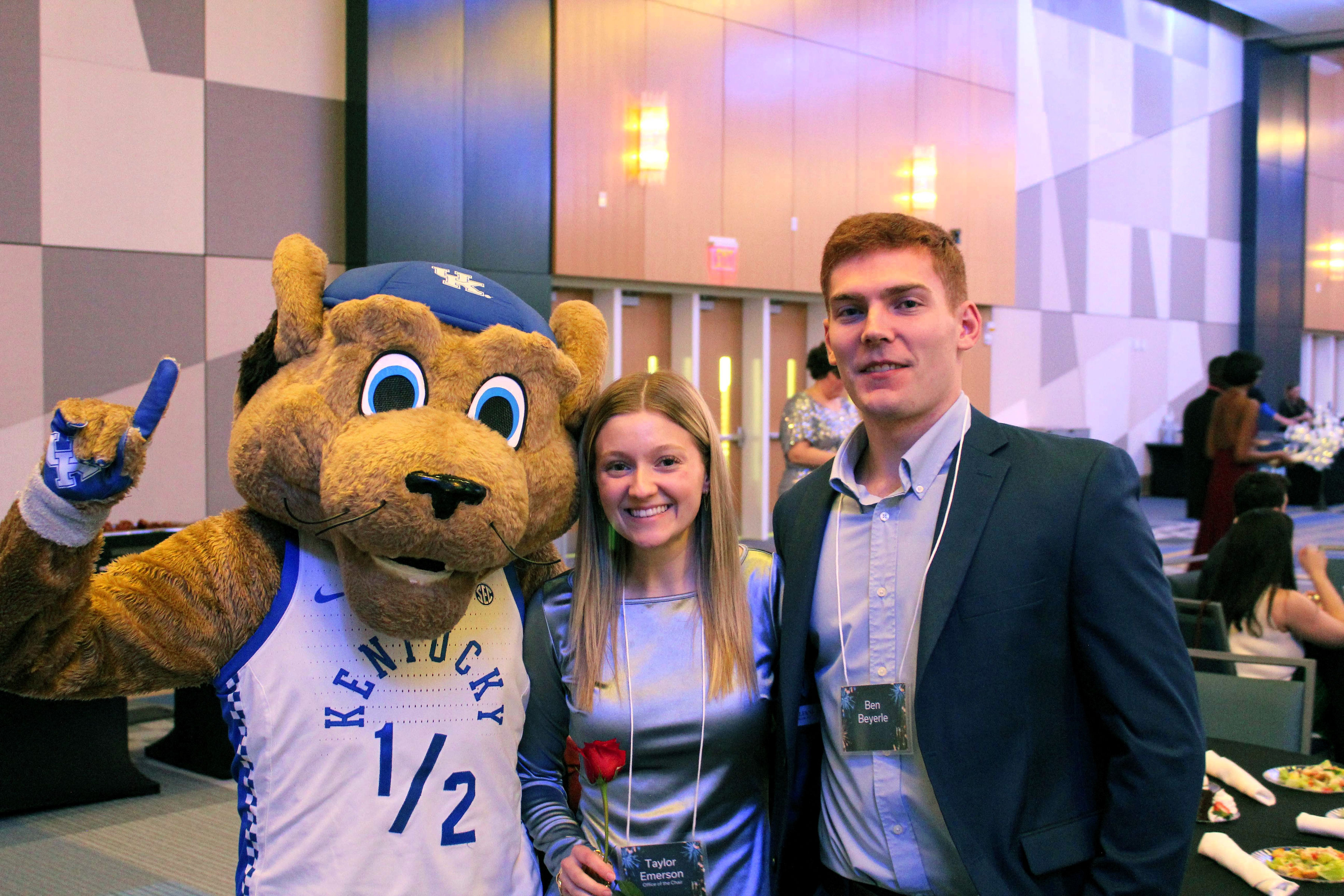 A young man and woman pose with Scratch the wildcat mascot. Scratch holds up a one with his fingers