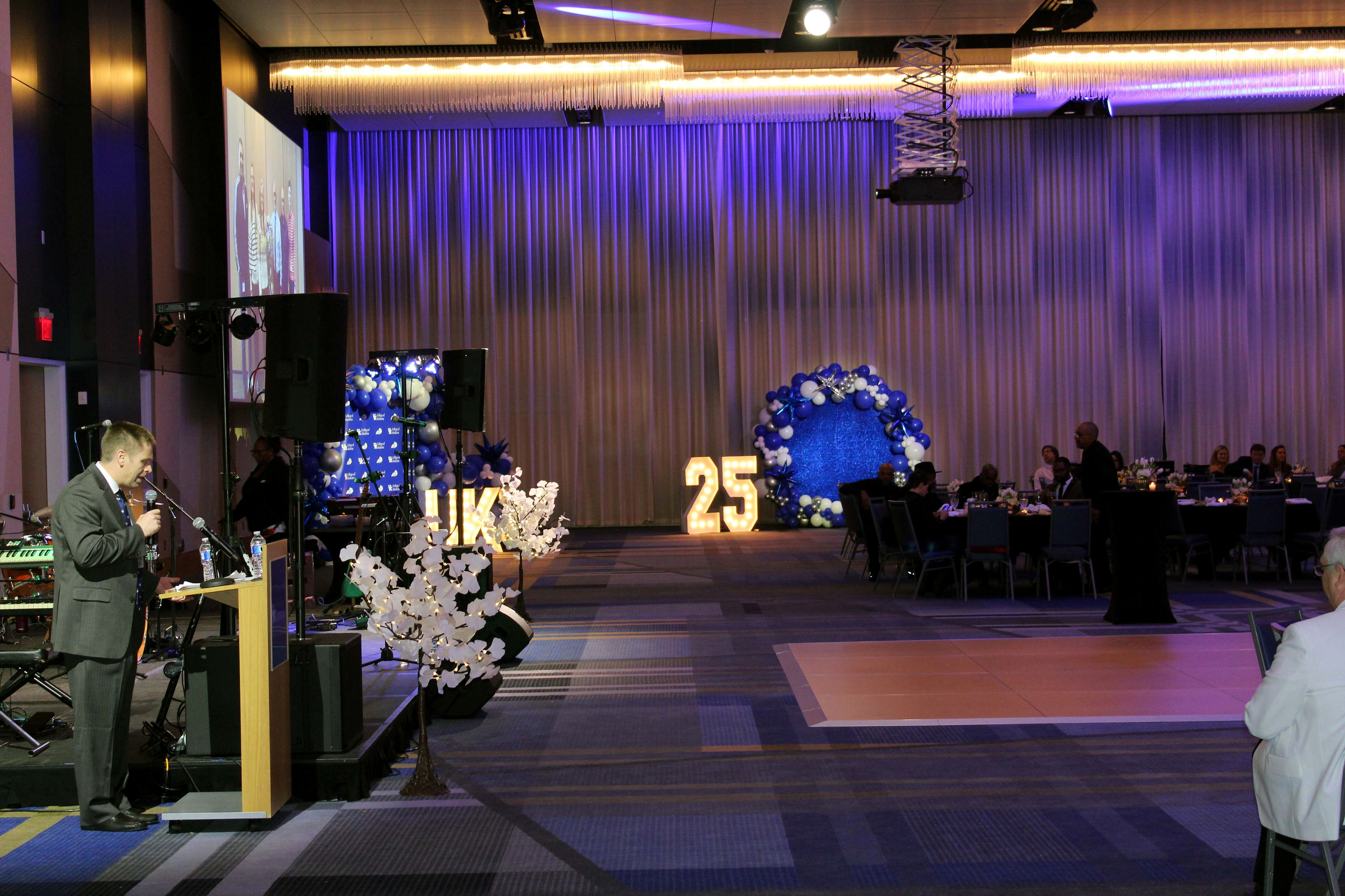 A speaker stands, haloed by a spotlight, in a fancy party hall. He holds a microphone. The crowd looks on