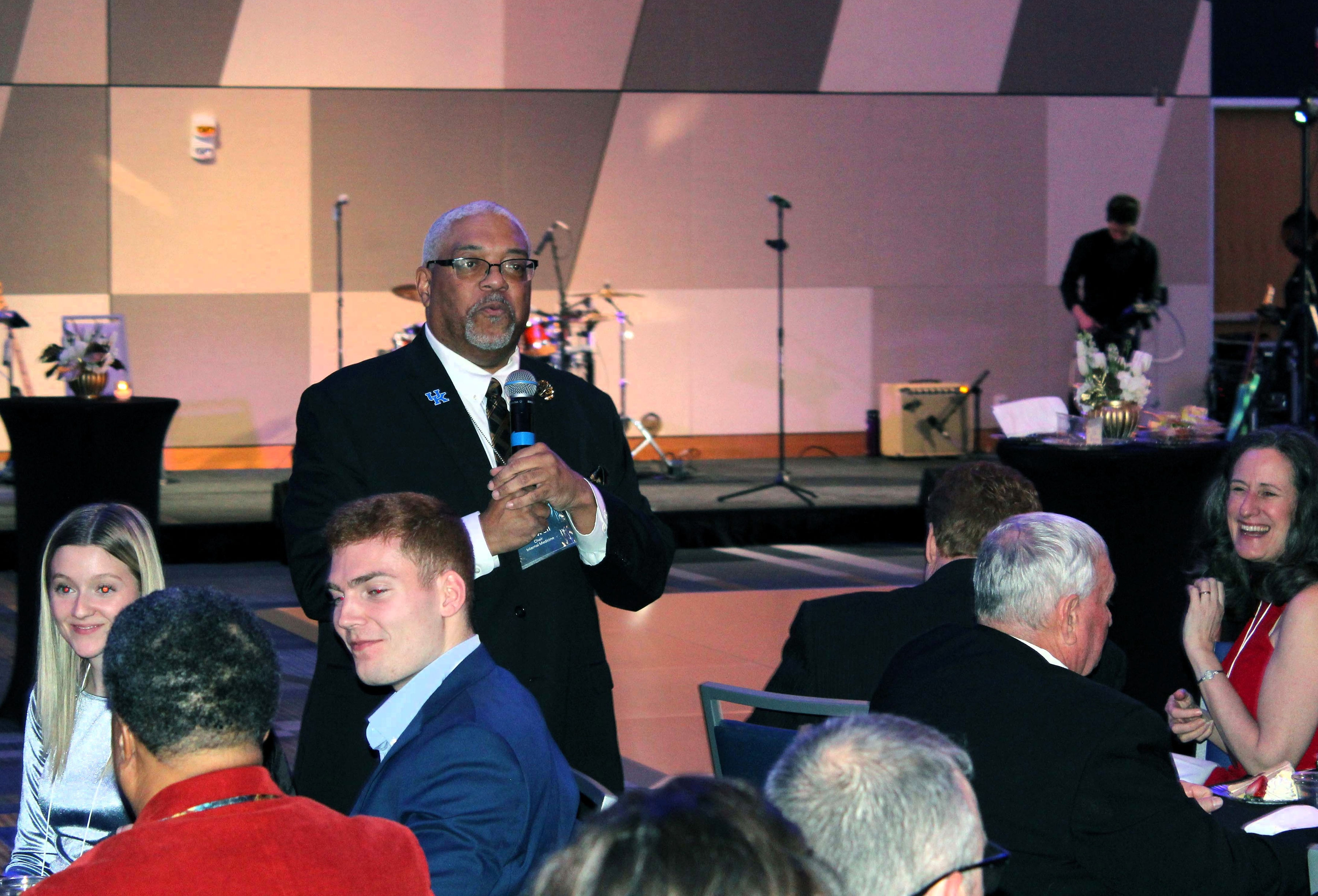 Dr. Conwell wears a suit and holds a microphone, and speaks to the room