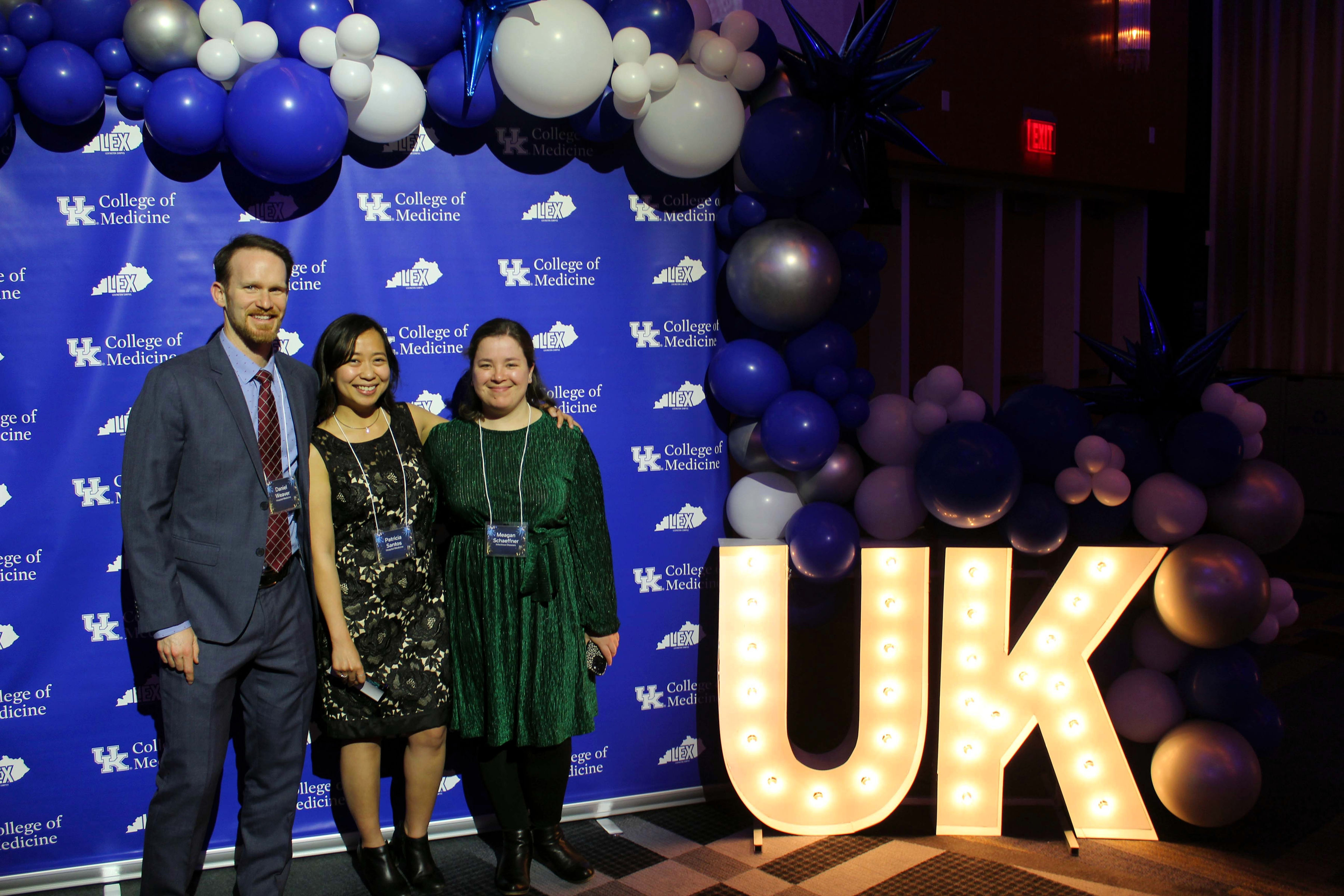 A man and two women in formal attire smile beneath a balloon arch. They stand next to large, glowing letters that say UK