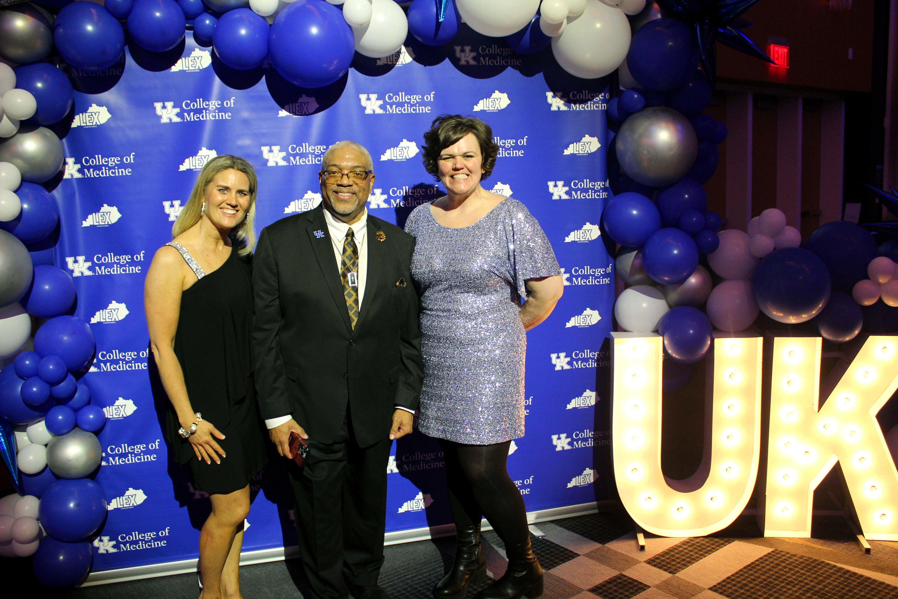 Dr. Conwell is flanked by two of his colleagues. They all smile beneath a balloon arch. Bright, glowing letters stand next to them that read "UK"