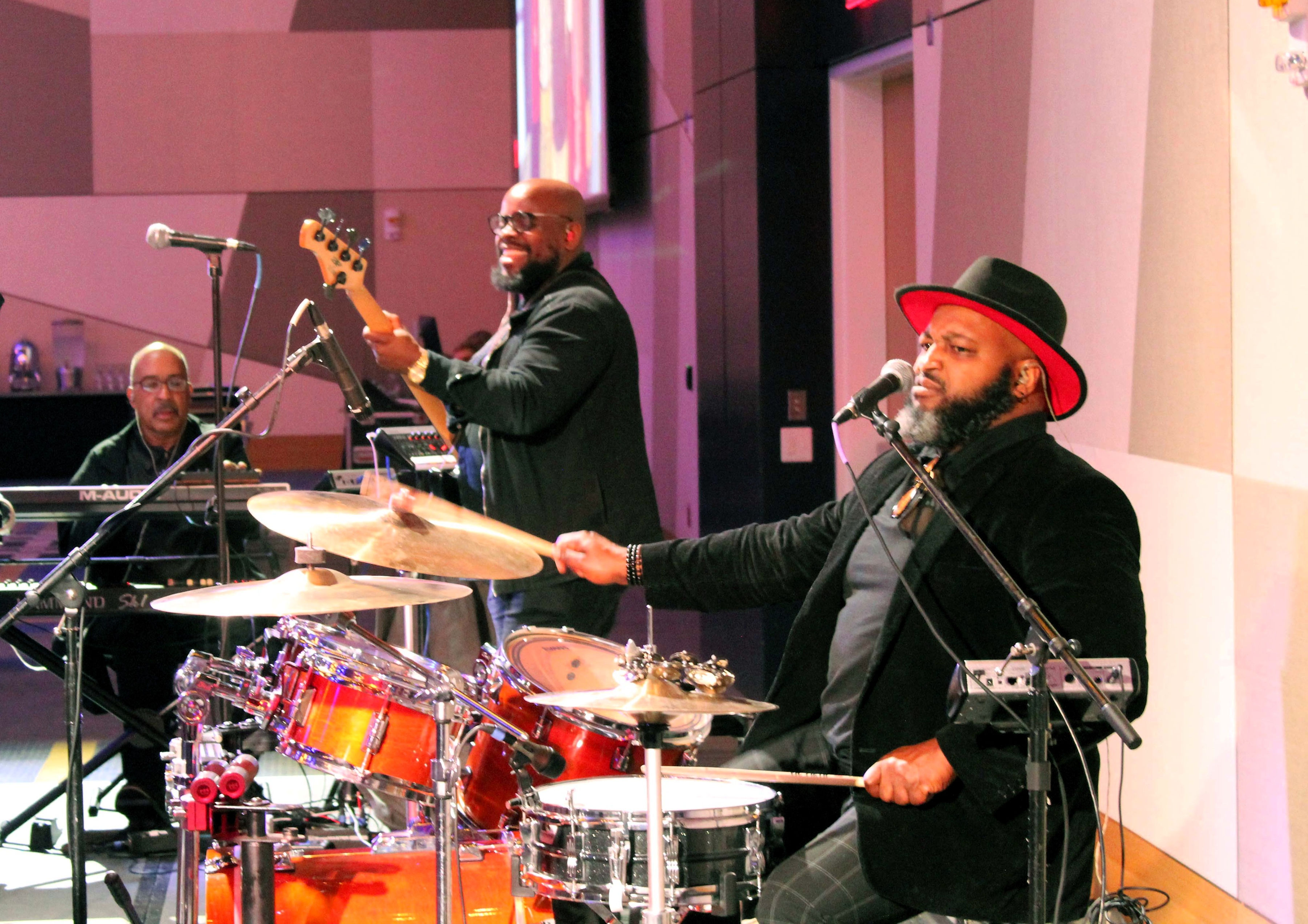 A band performs on a stage amidst sound recording equipment. The drummer is playing with a focused look. The guitarist is smiling