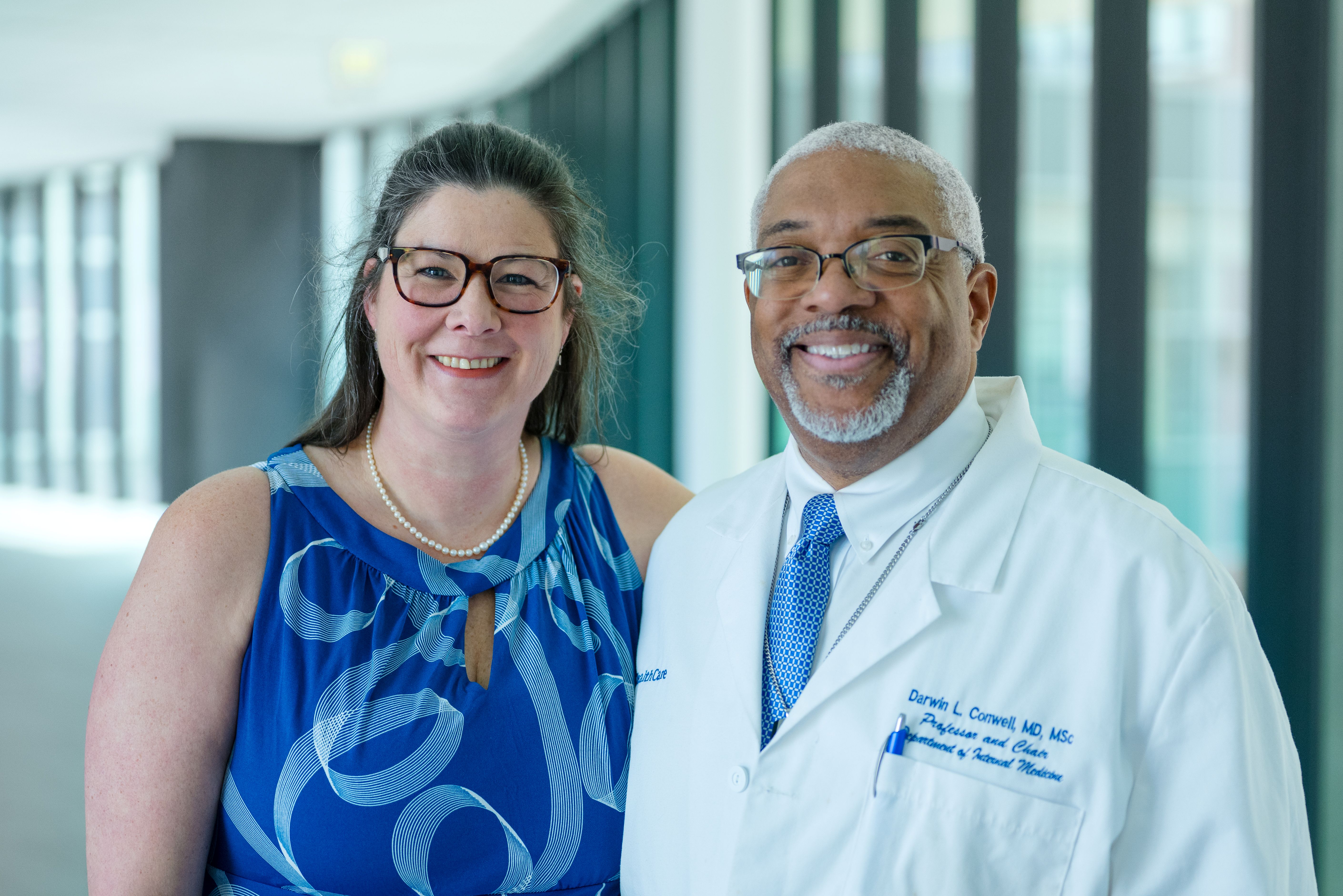 A man and a woman smile at the camera. The man wears a lab coat, and the woman wears a blue dress