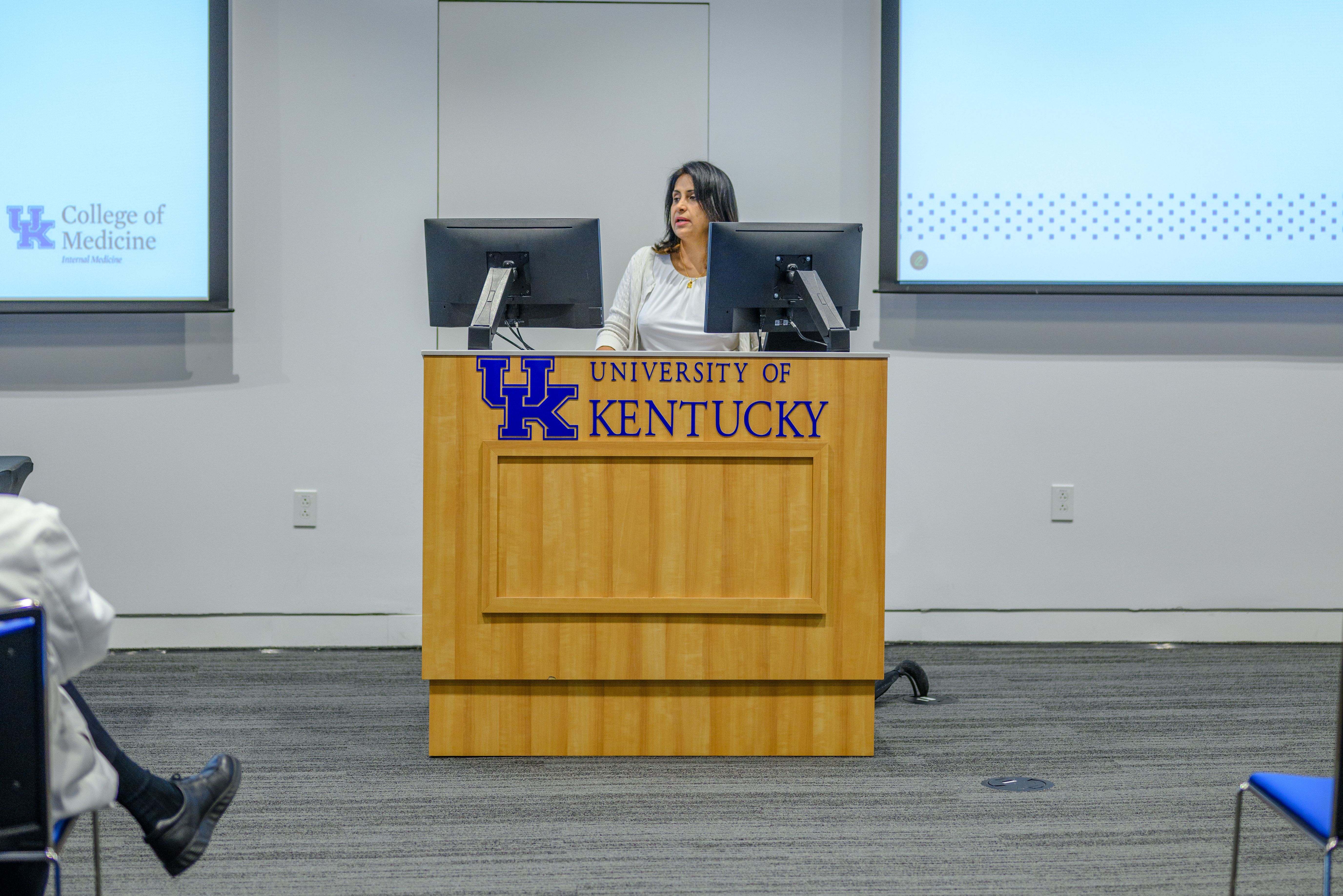 A woman speaks at a podium. The podium says, "University of Kentucky" on the front