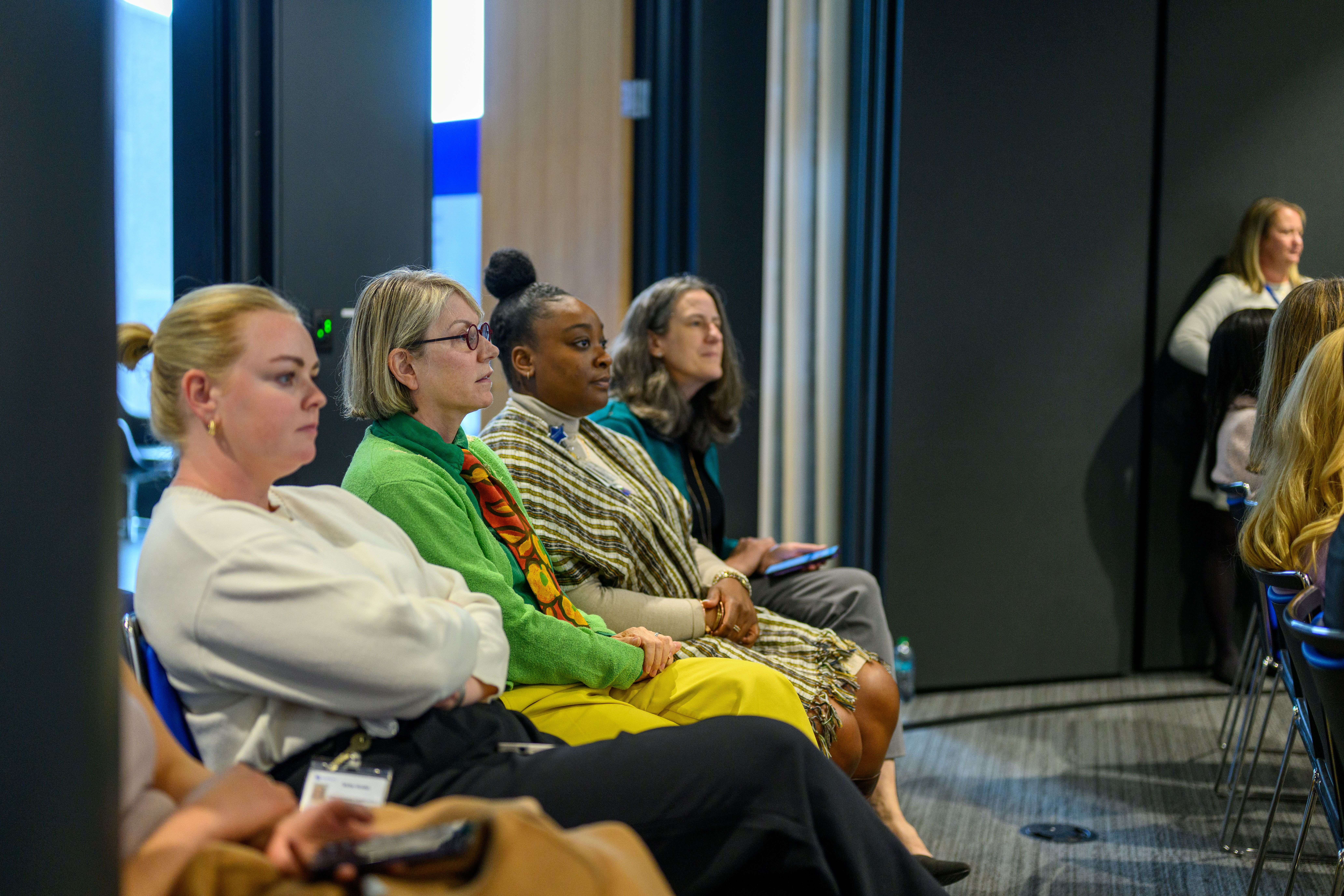 Four women sit in a line watching a presentation off screen. They are all focused and interested.