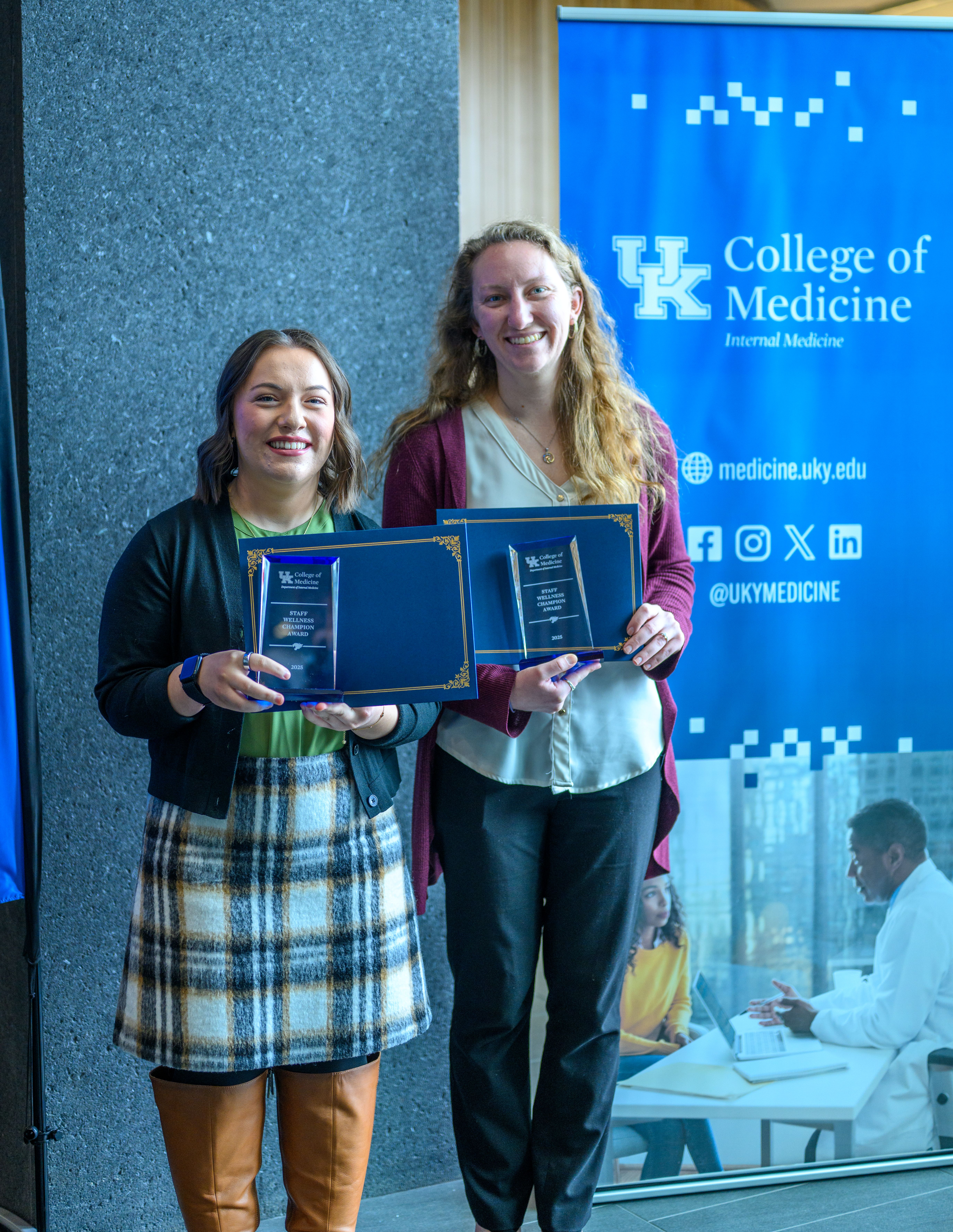 Two women hold up glass awards, smiling. They stand in front of a blue banner that says College of Medicine; Internal Medicine on it