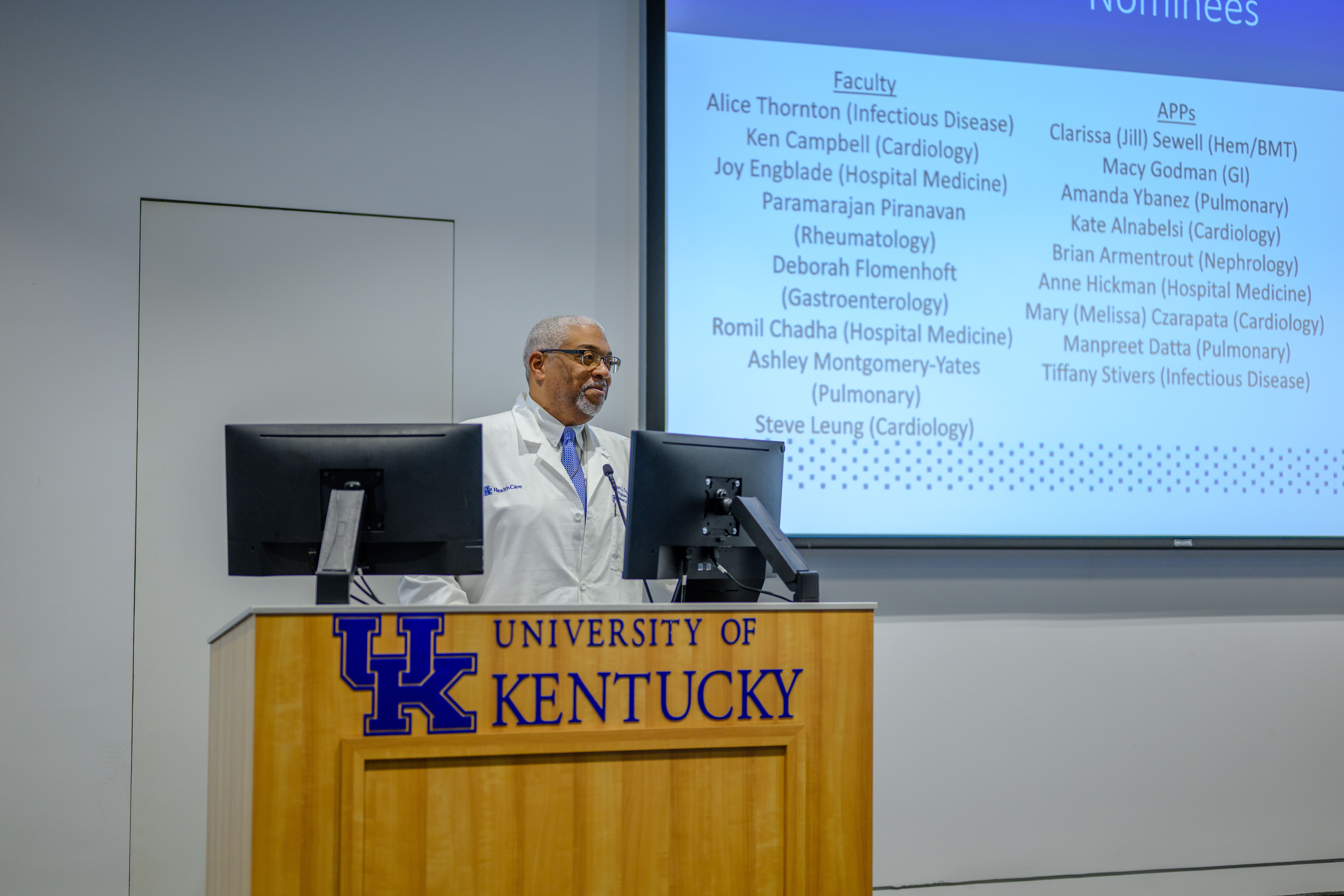 A man in a lab coat stands at a podium. The podium says "University of Kentucky." Behind him on a screen, nominees for an award are listed