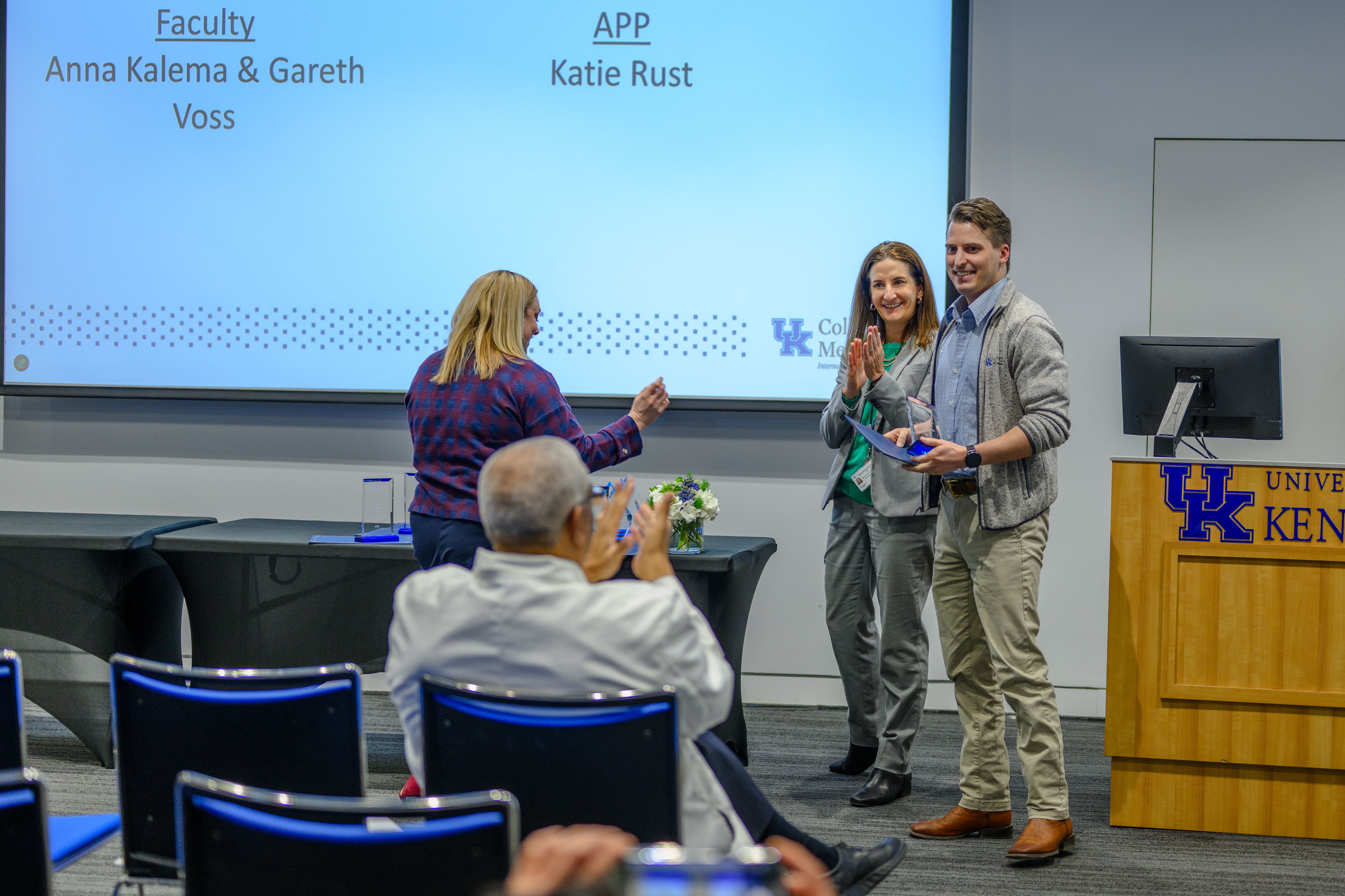 A man stands with a glass award. Around him, three people clap