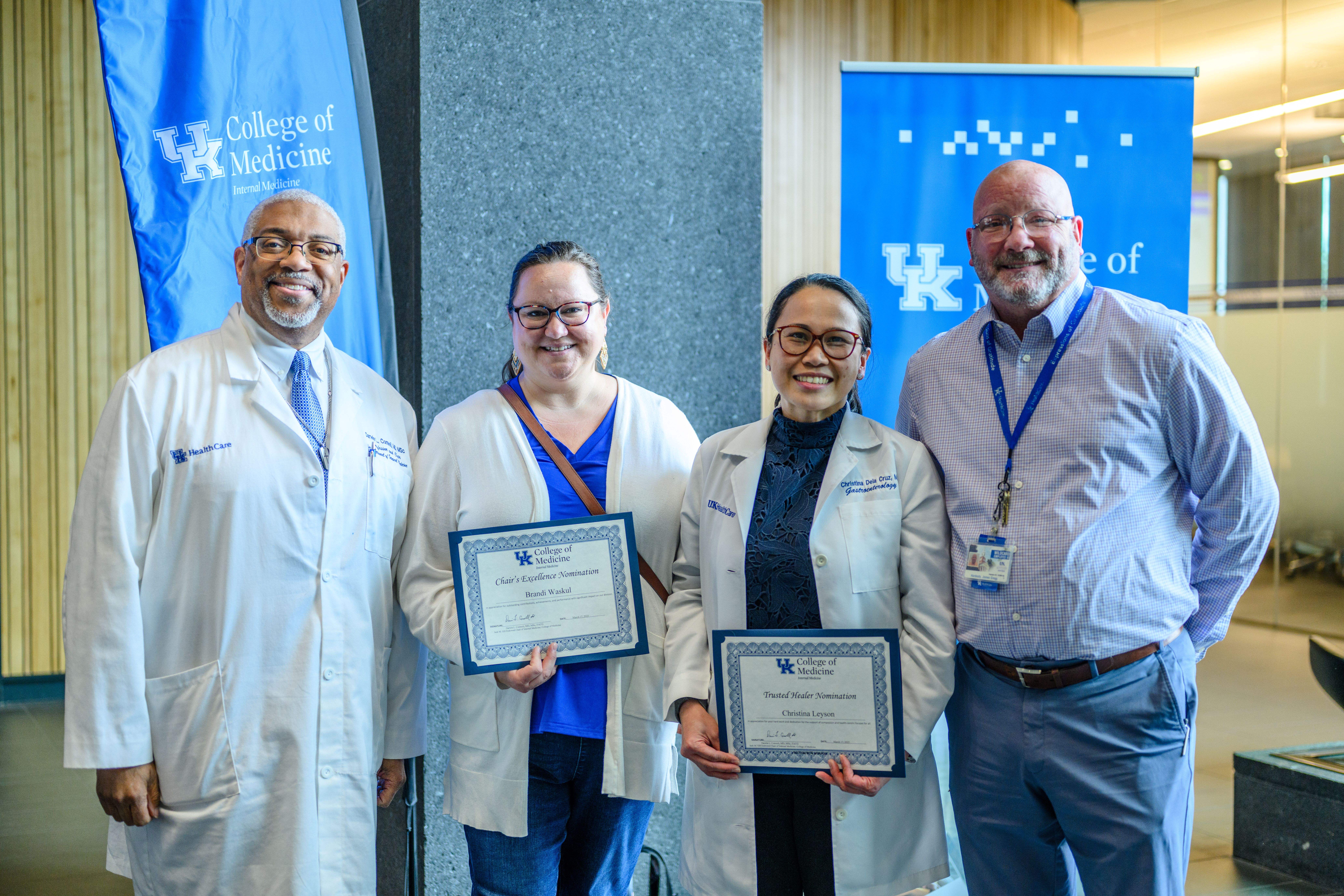 Four people stand in a row. The two women in the middle hold up certificates. They are all smiling