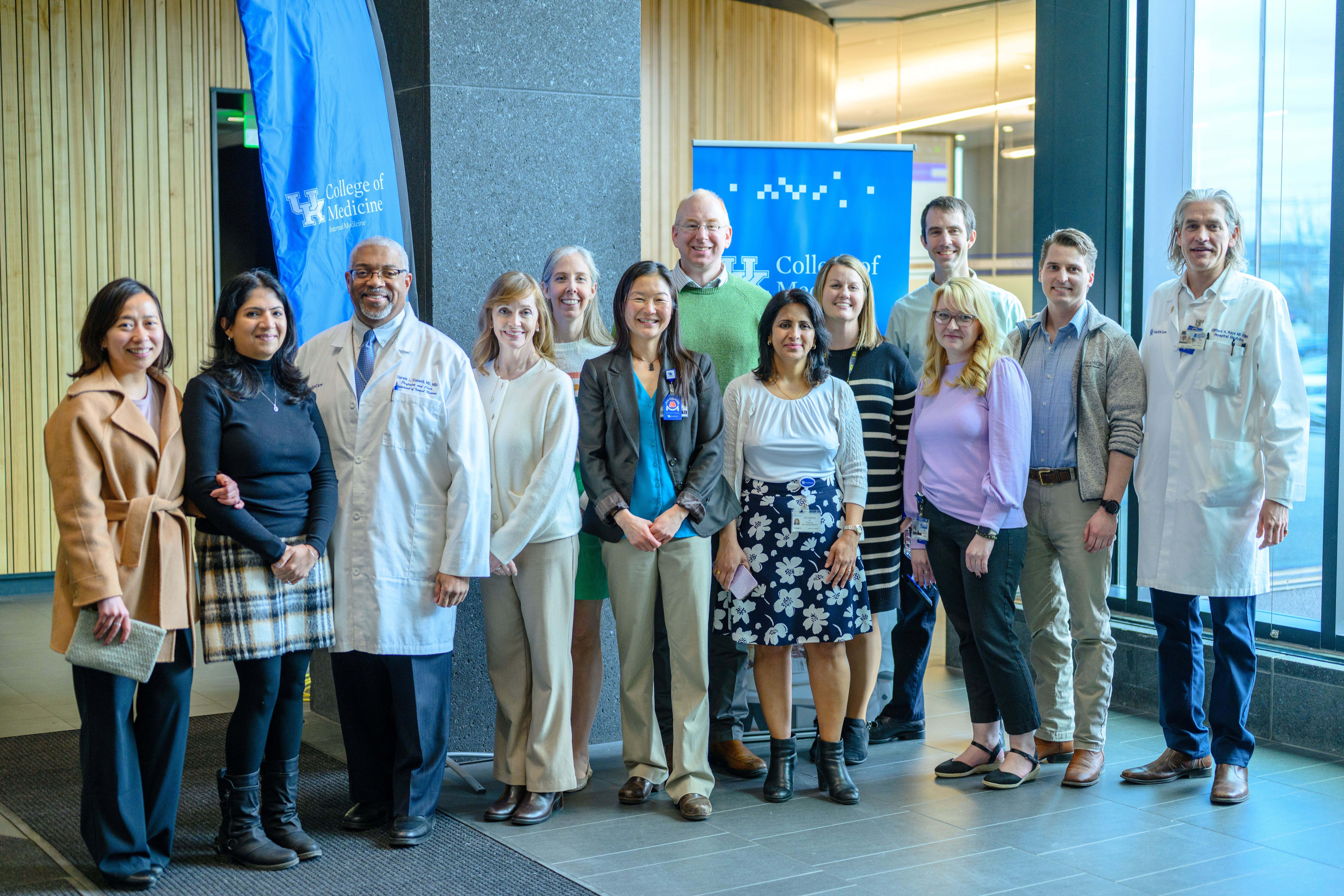 A large group of people pose for a photo. They are all standing in nice business clothes. Two of them wear lab coats.