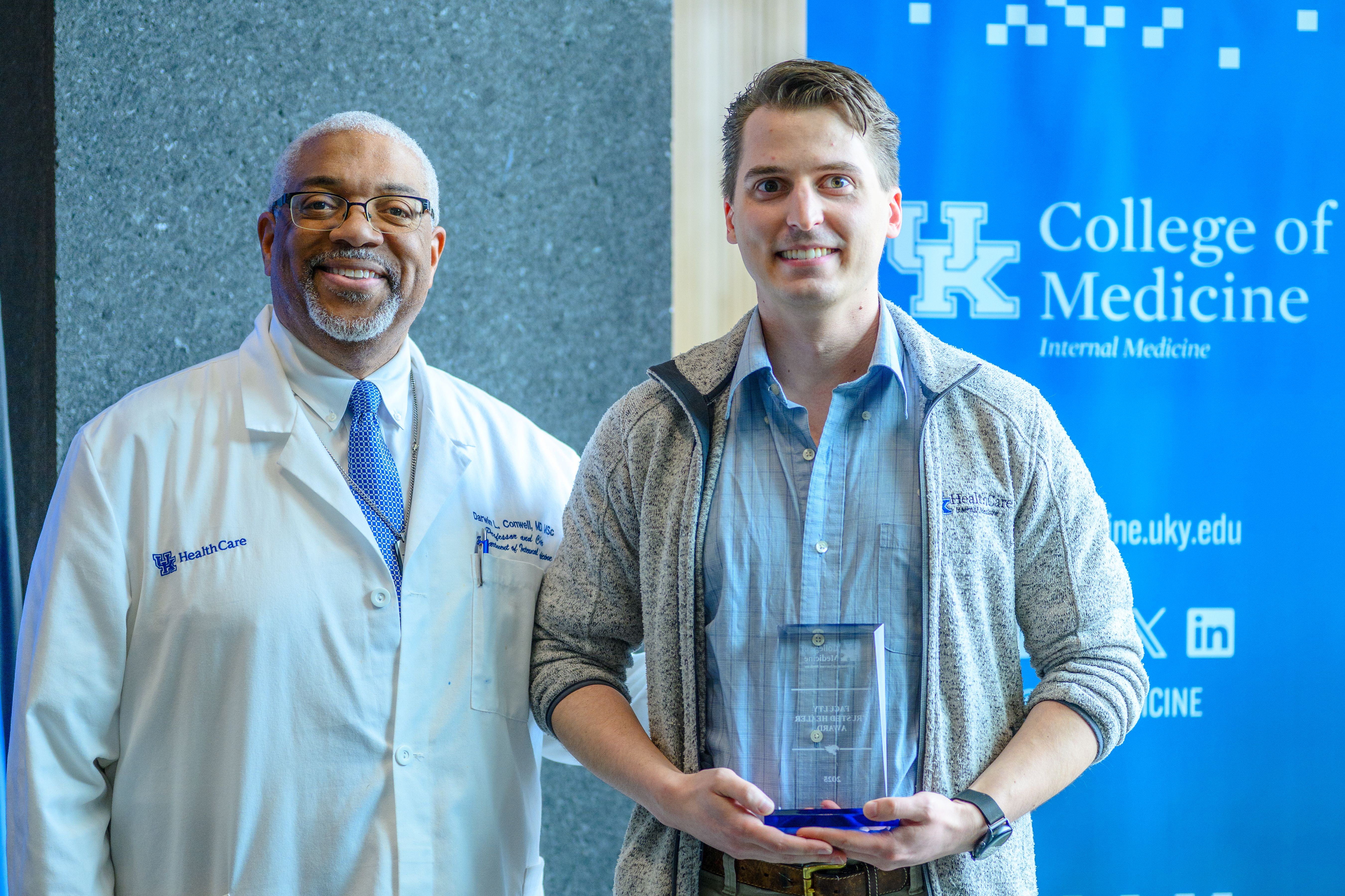 Two men stand next to eac h other, smiling. One wears a lab coat. The other holds a glass award.
