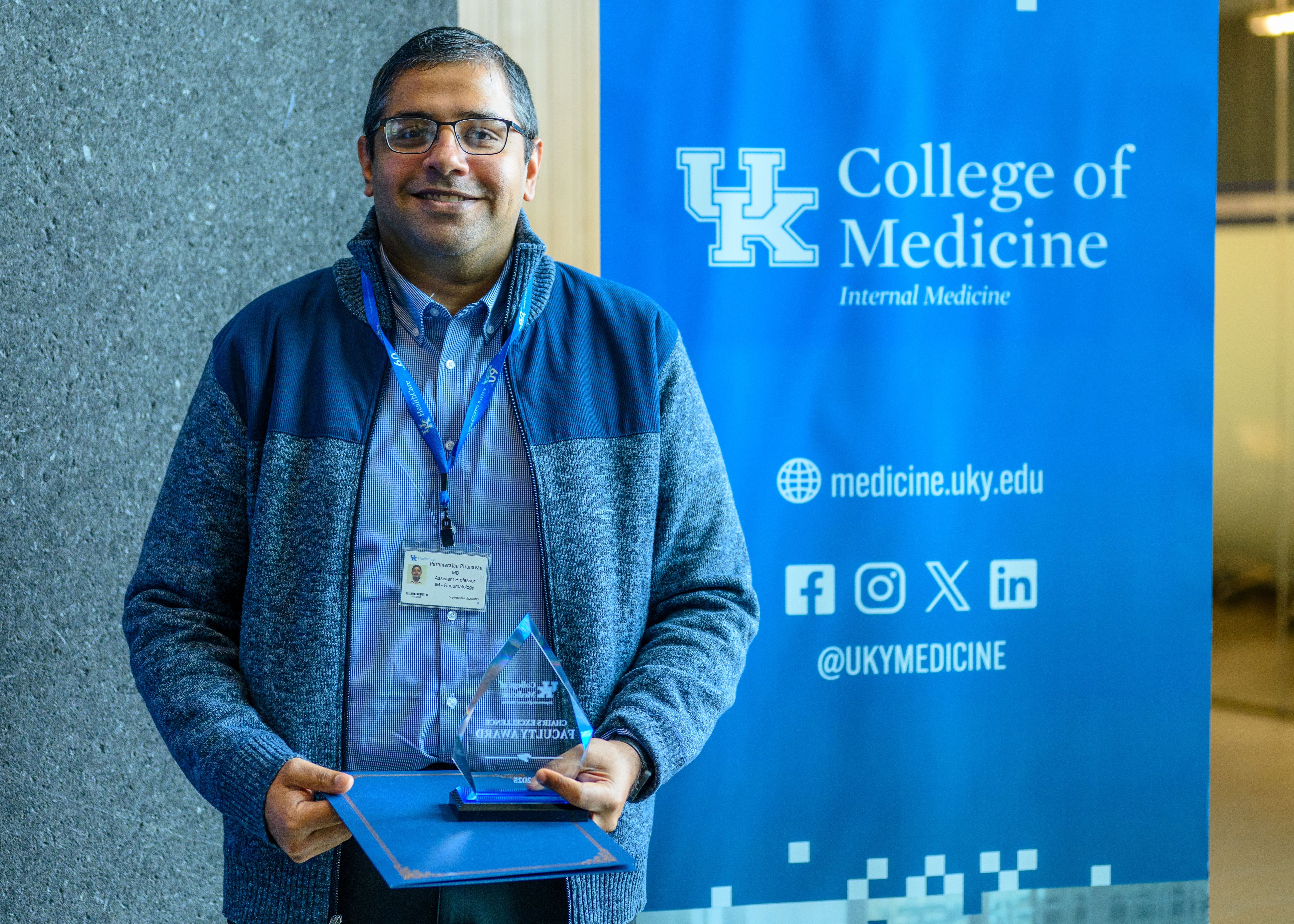 A man stands in front of a blue banner that says "College of Medicine; Internal Medicine." He is holding a glass award and smiling