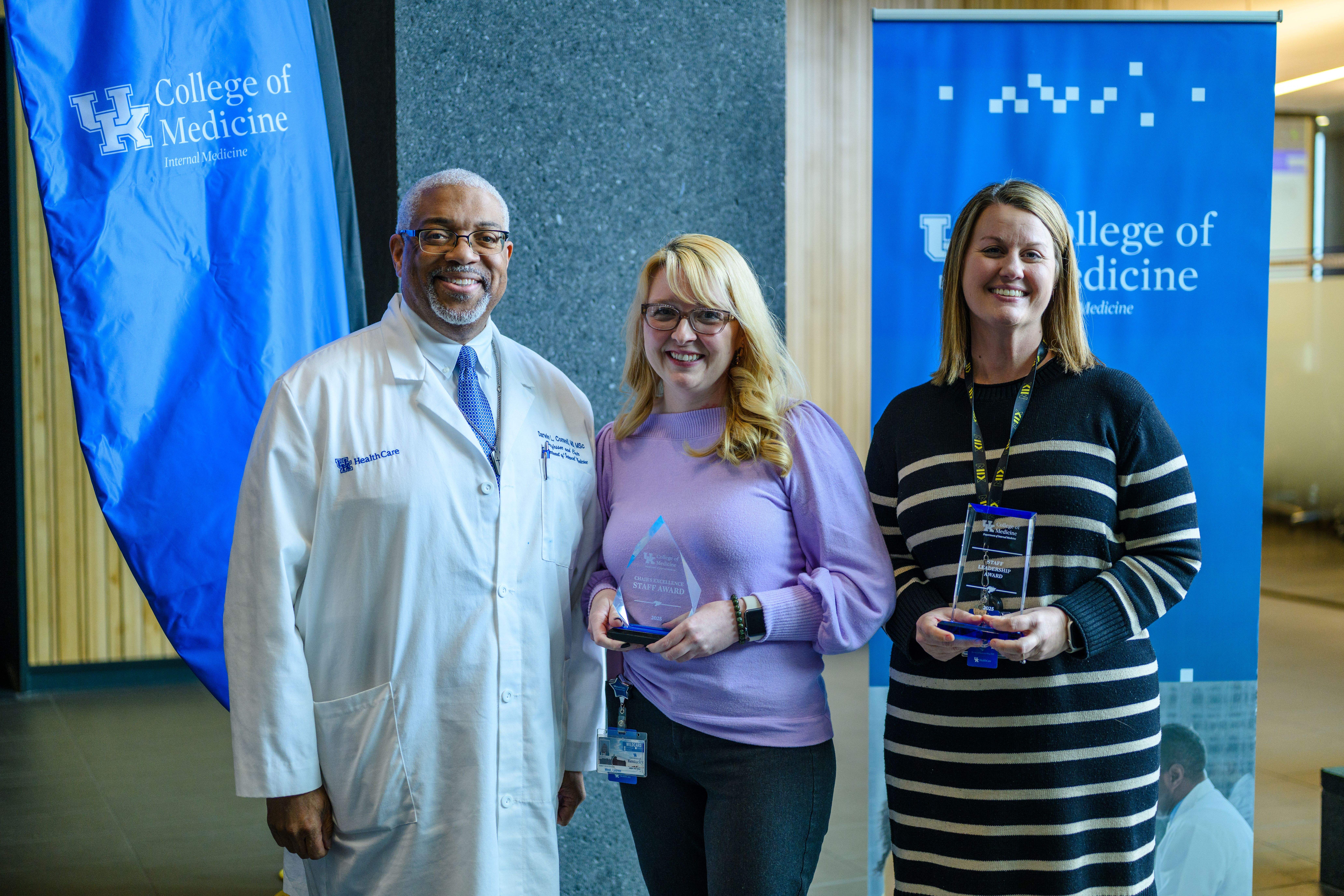 Two women stand smiling, holding glass awards. To the left, a man in a lab coat smiles as well