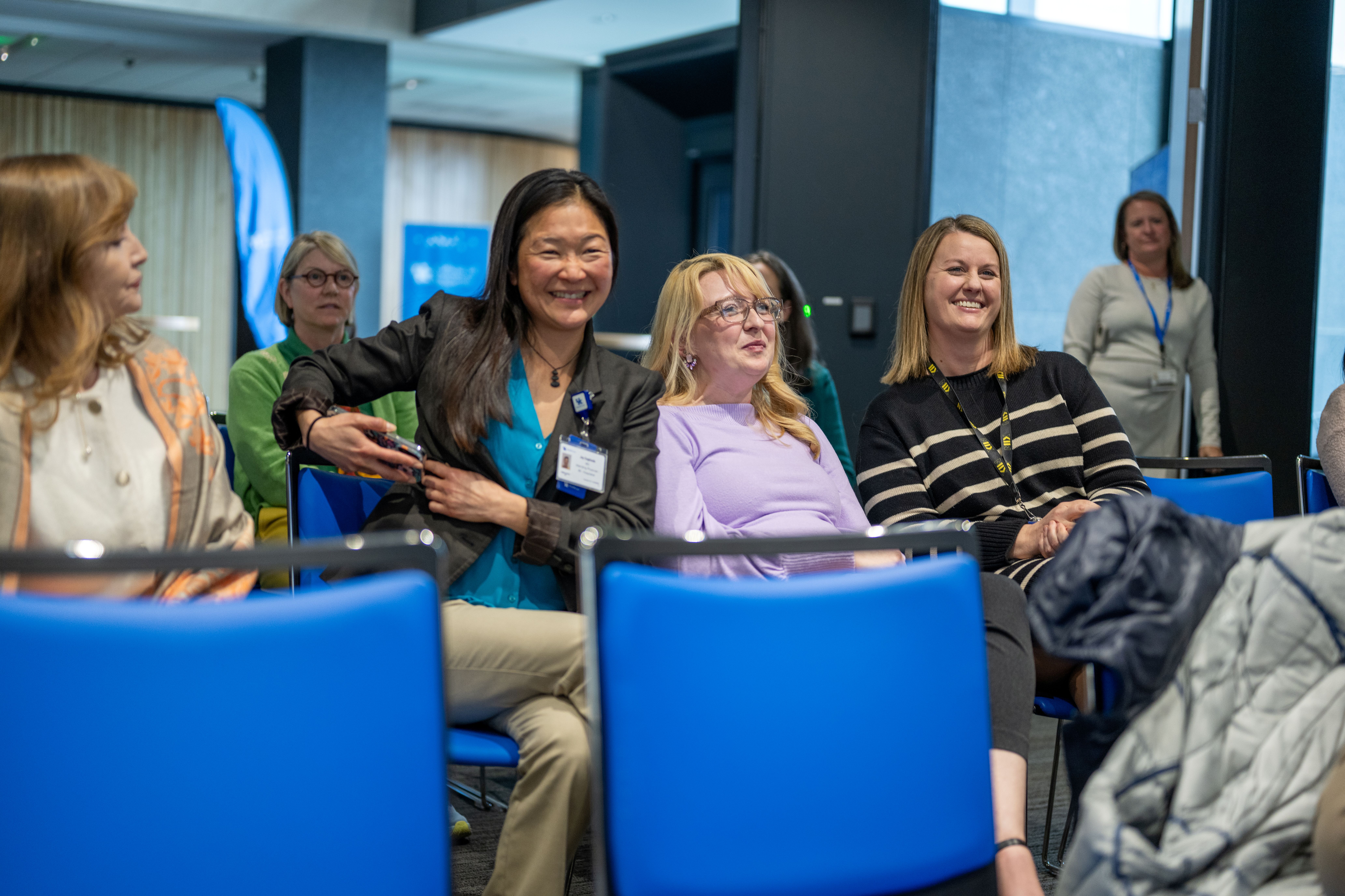A group of women sit in a row of chairs, watching a presentation off screen. The woman in the center of the frame is smiling very big.