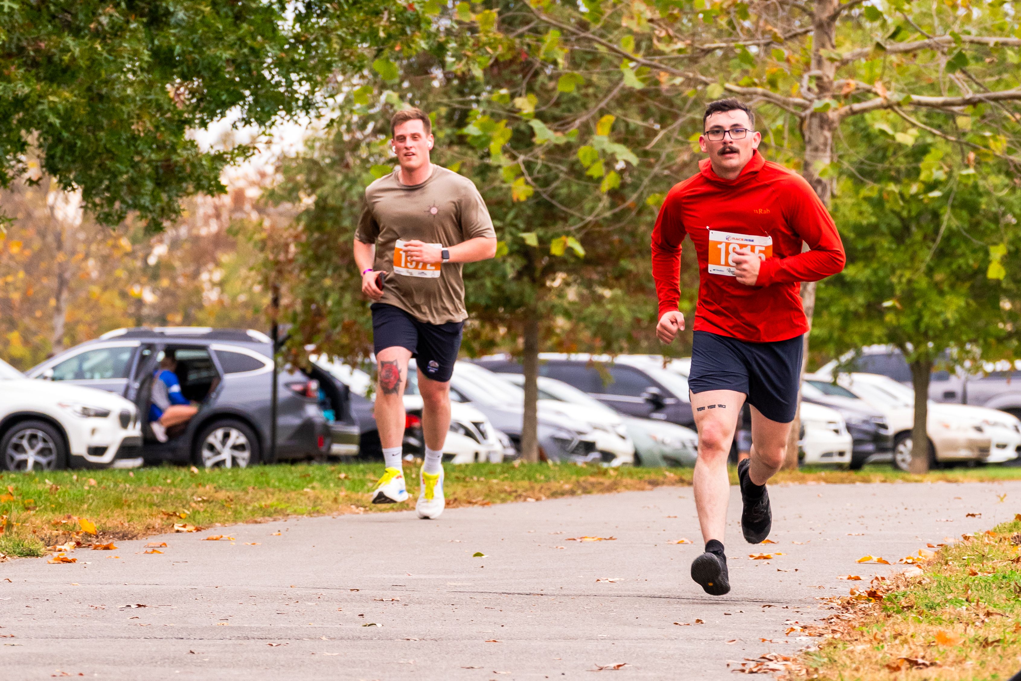 Two men run on a concrete path beneath Autum trees. They look focused and happy.