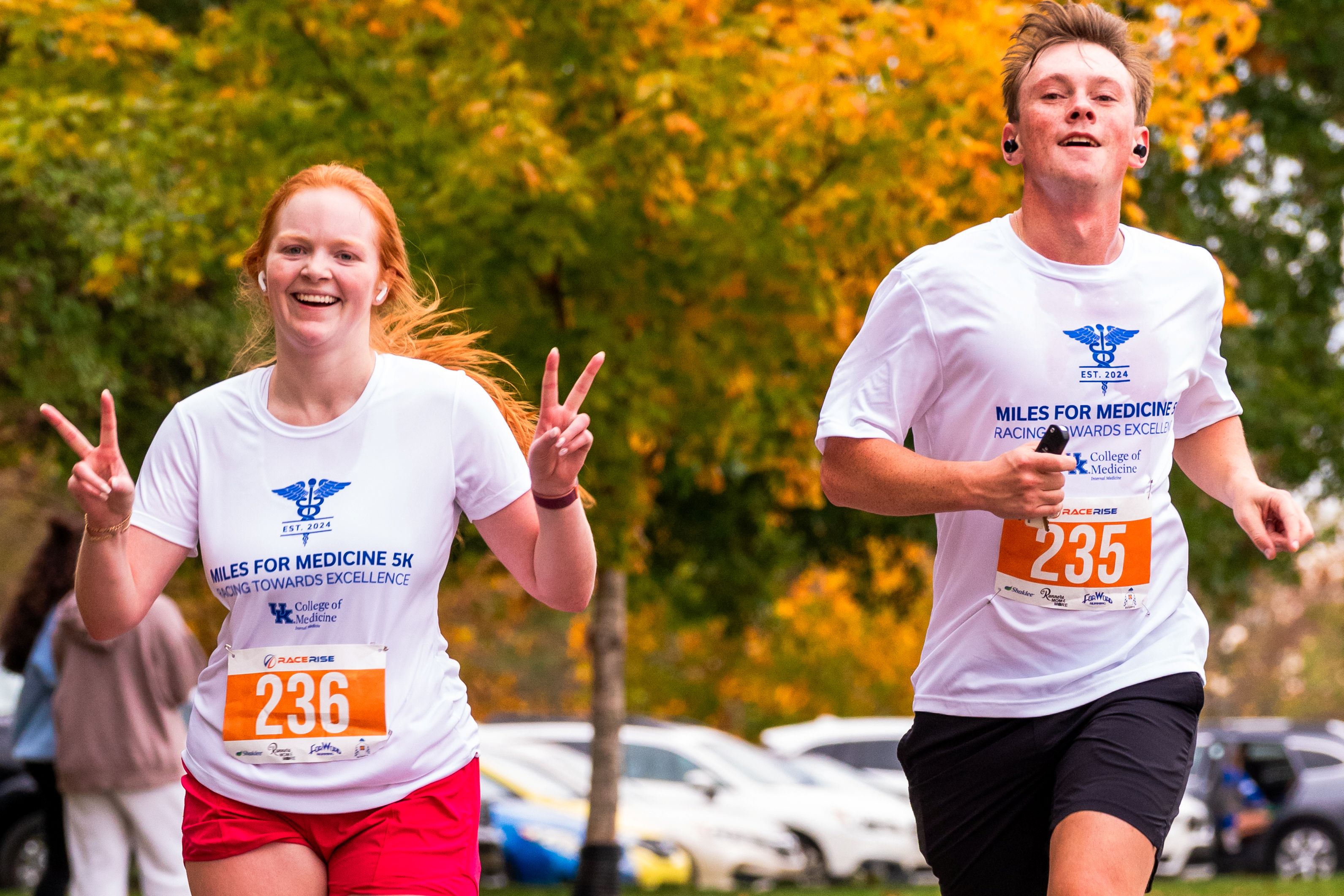 A man and a woman run beneath Autumn trees in white shirts that say "Miles for Medicine." The woman is holding up a double peace sign.