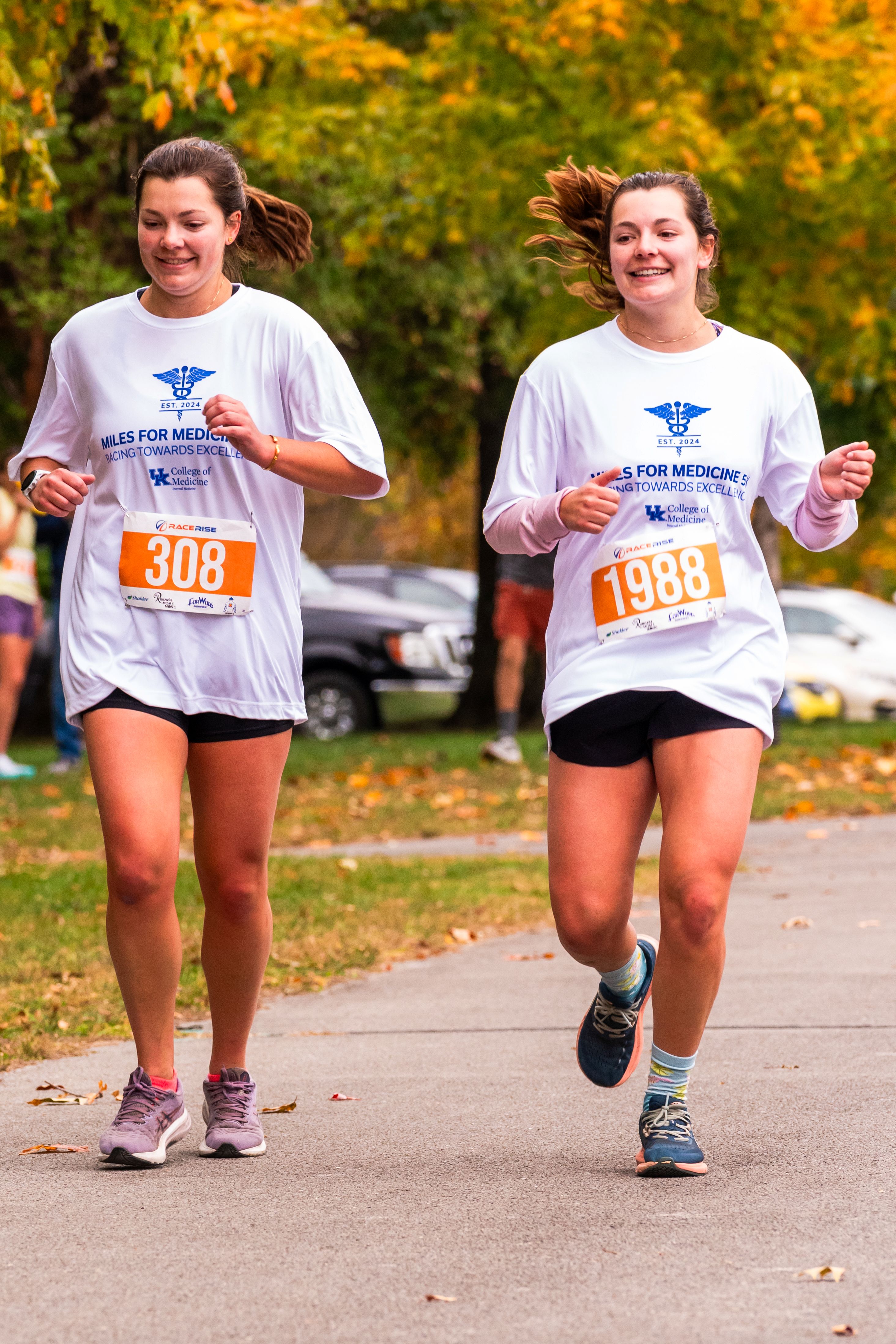 Two women with ponytails run in white shirts that say "Miles for Medicine." They are both smiling.