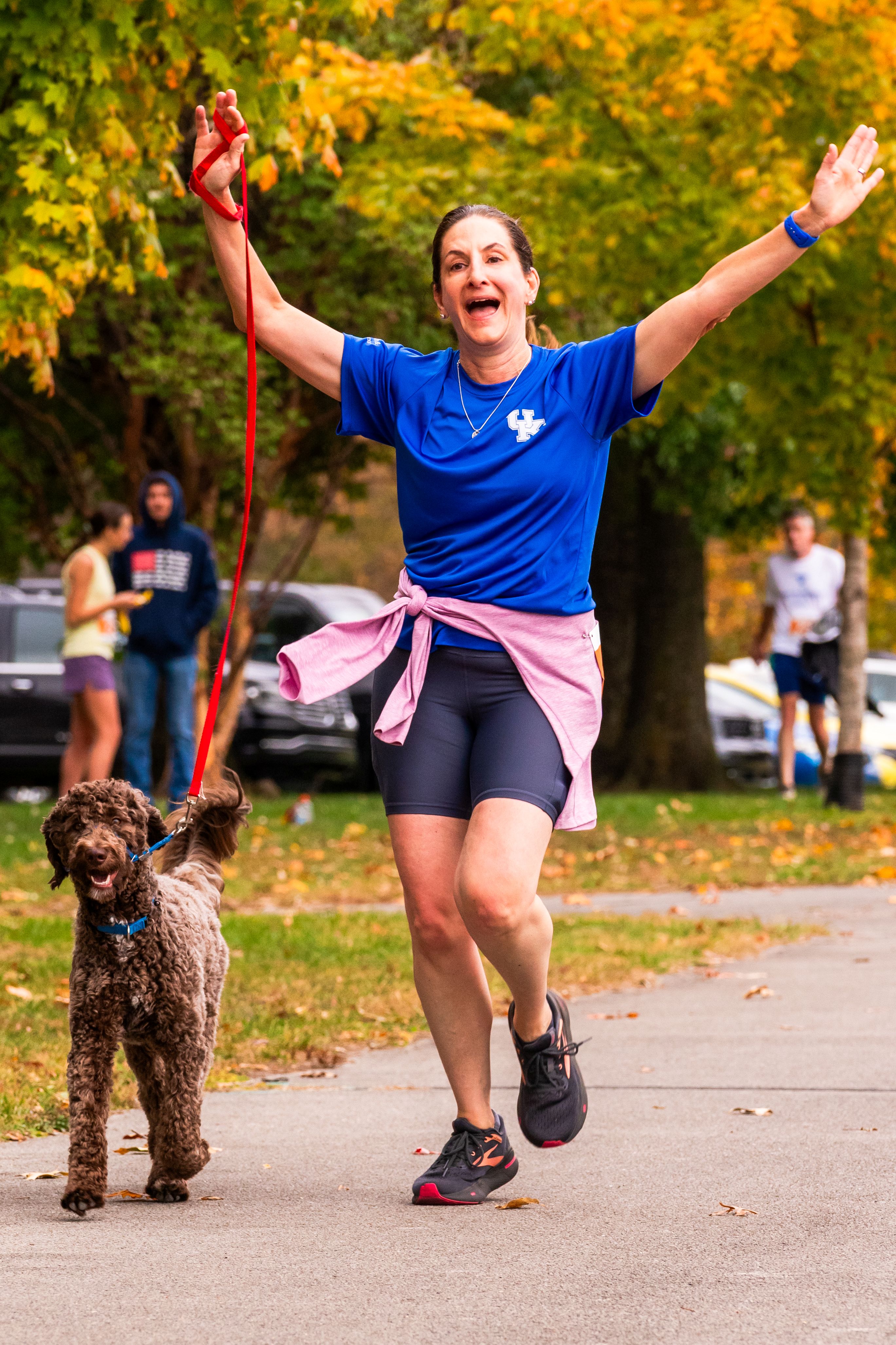 A woman in a blue shirt holds her arms up in a V as she crosses the finish line. She runs with a brown dog on a leash