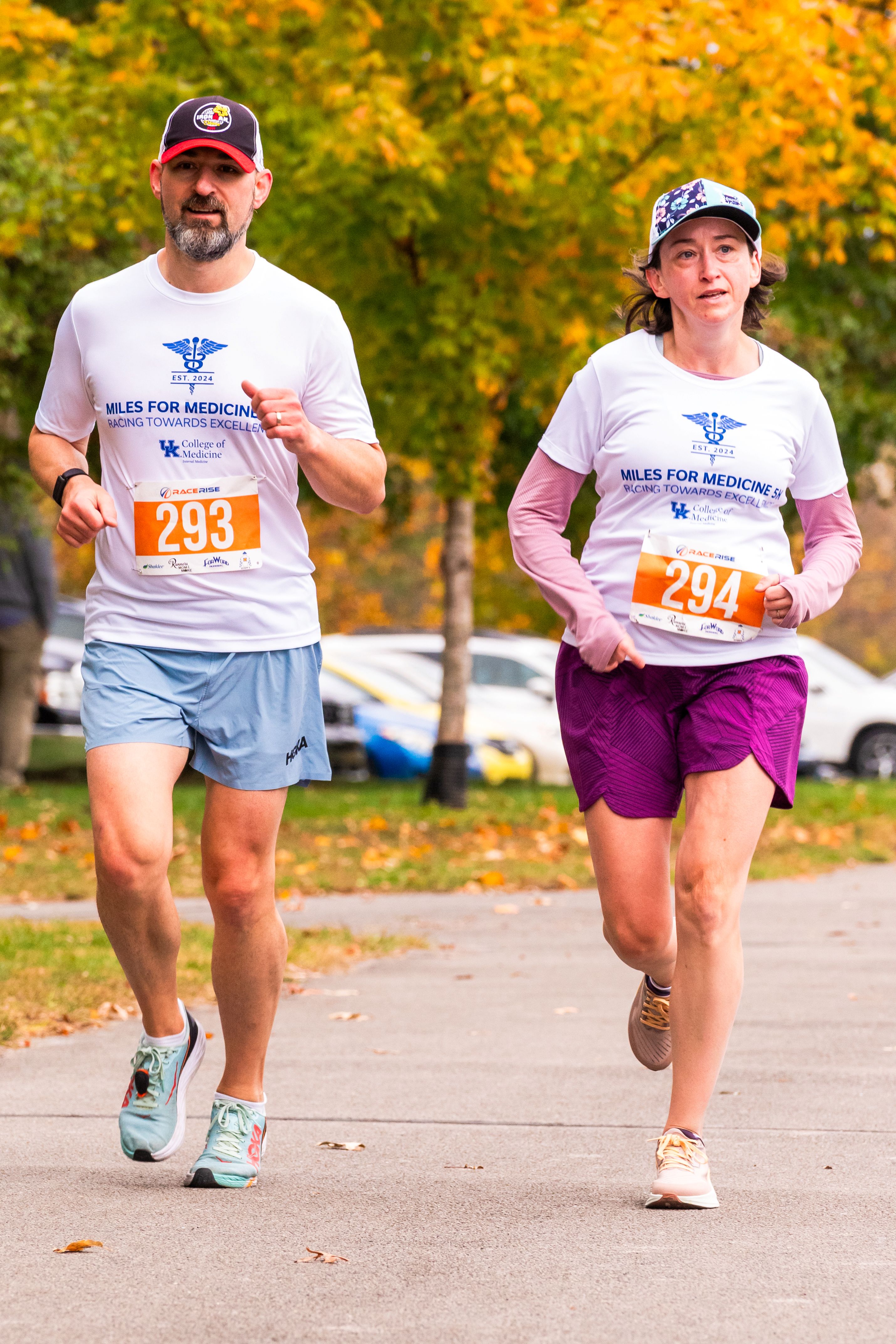 A man and woman jog lightly with focused looks. They both wear white shirts that say "Miles for Medicine"