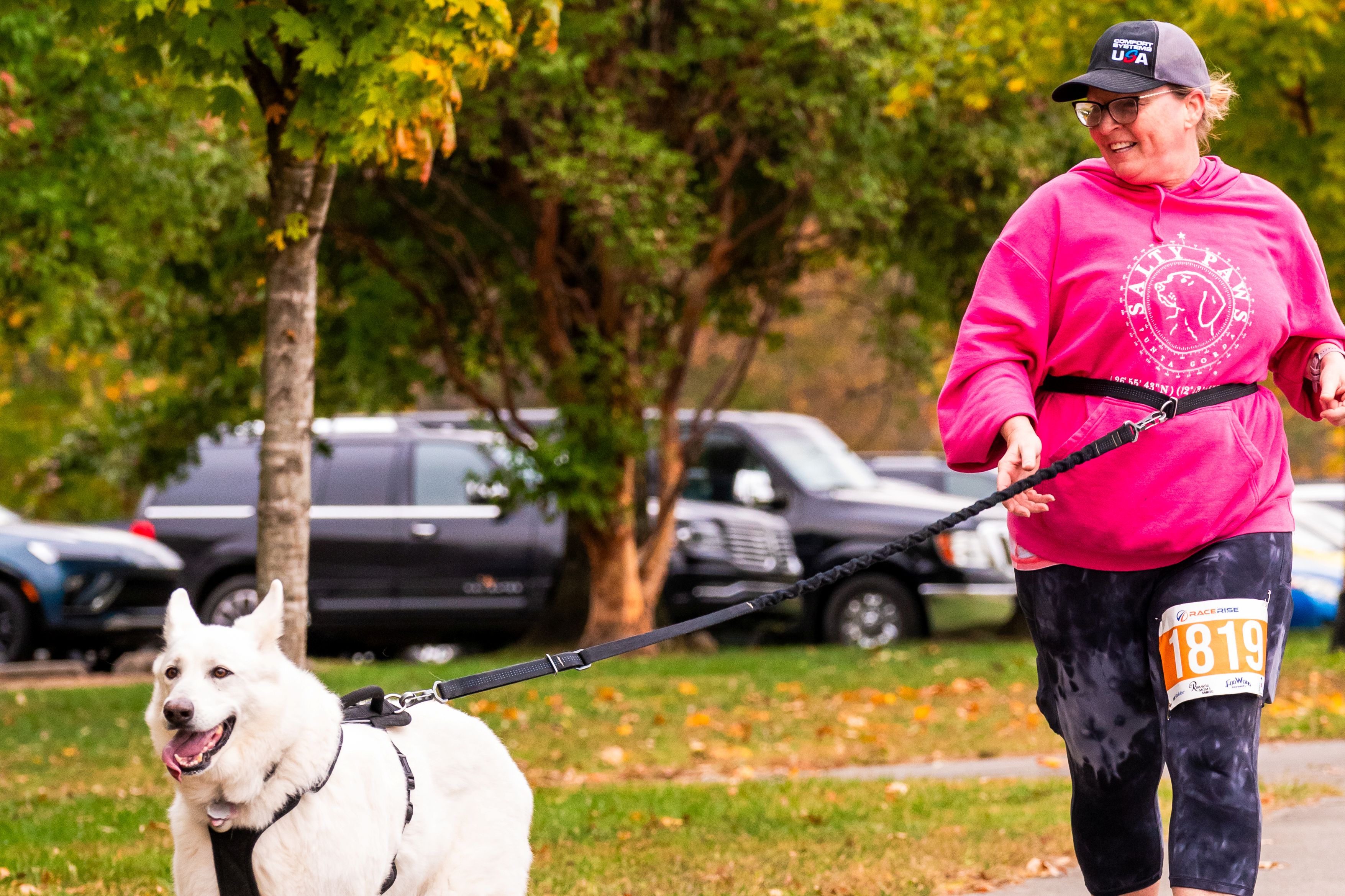 A woman holding a leash runs while smiling. Her large, white dog runs in front happily.