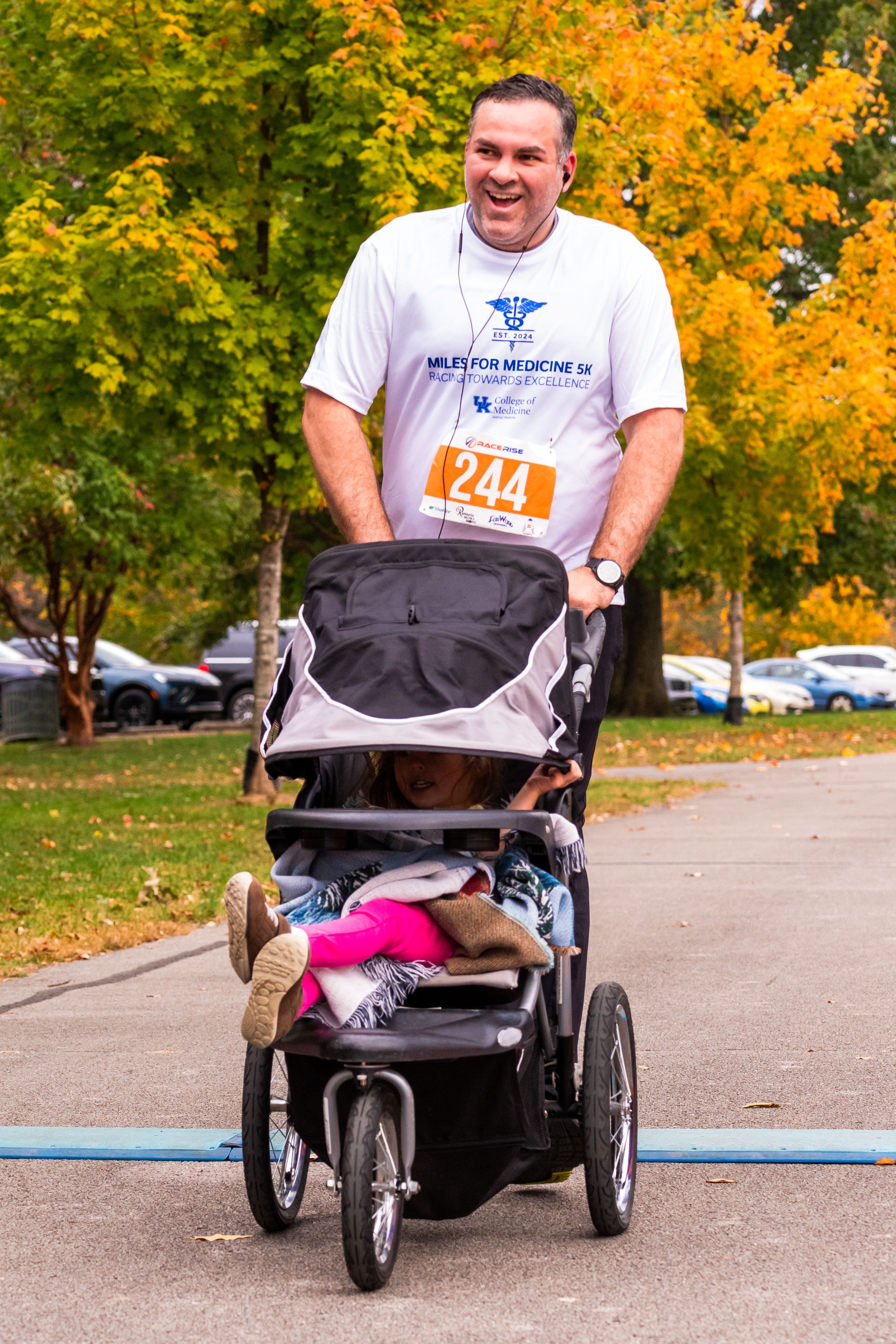 A man runs while pushing a stroller. He wears a white shirt that says "Miles for Medicine"