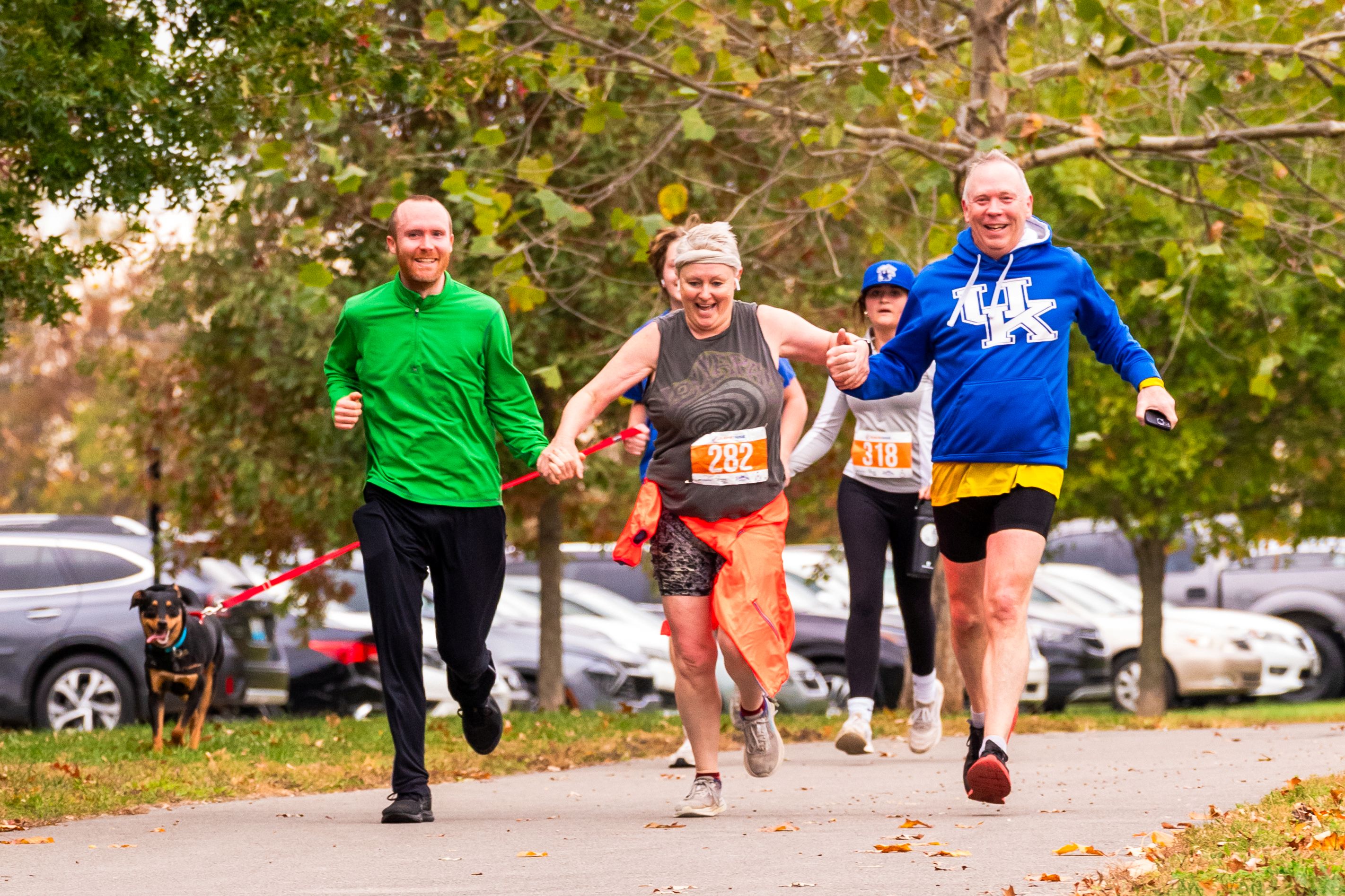 Two men and a woman run while holding hands. The woman is in the middle. They are all smiling.