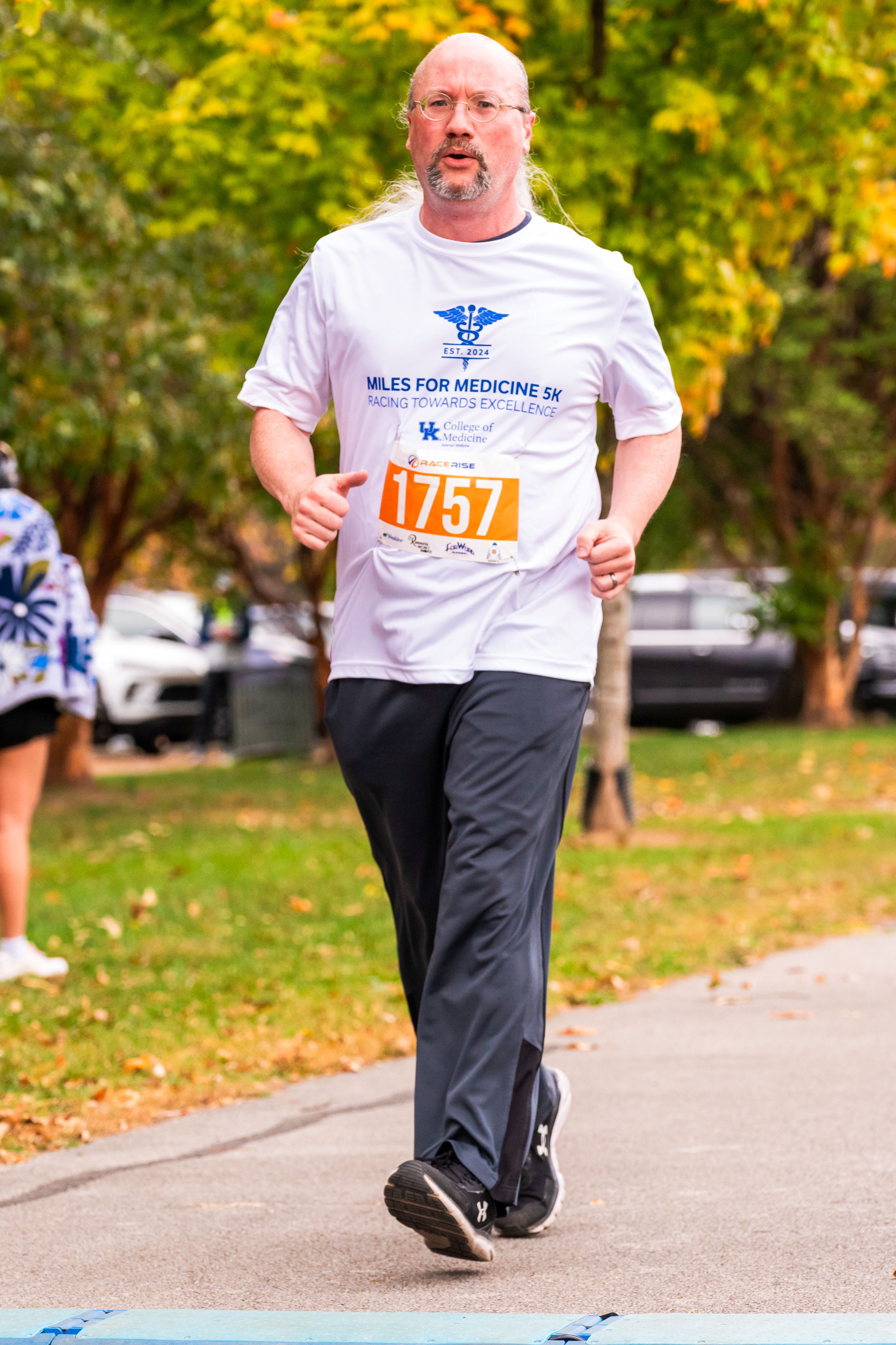 A man with glasses jogs with a focused look on his face. He wears a white shirt that says "Miles for Medicine"