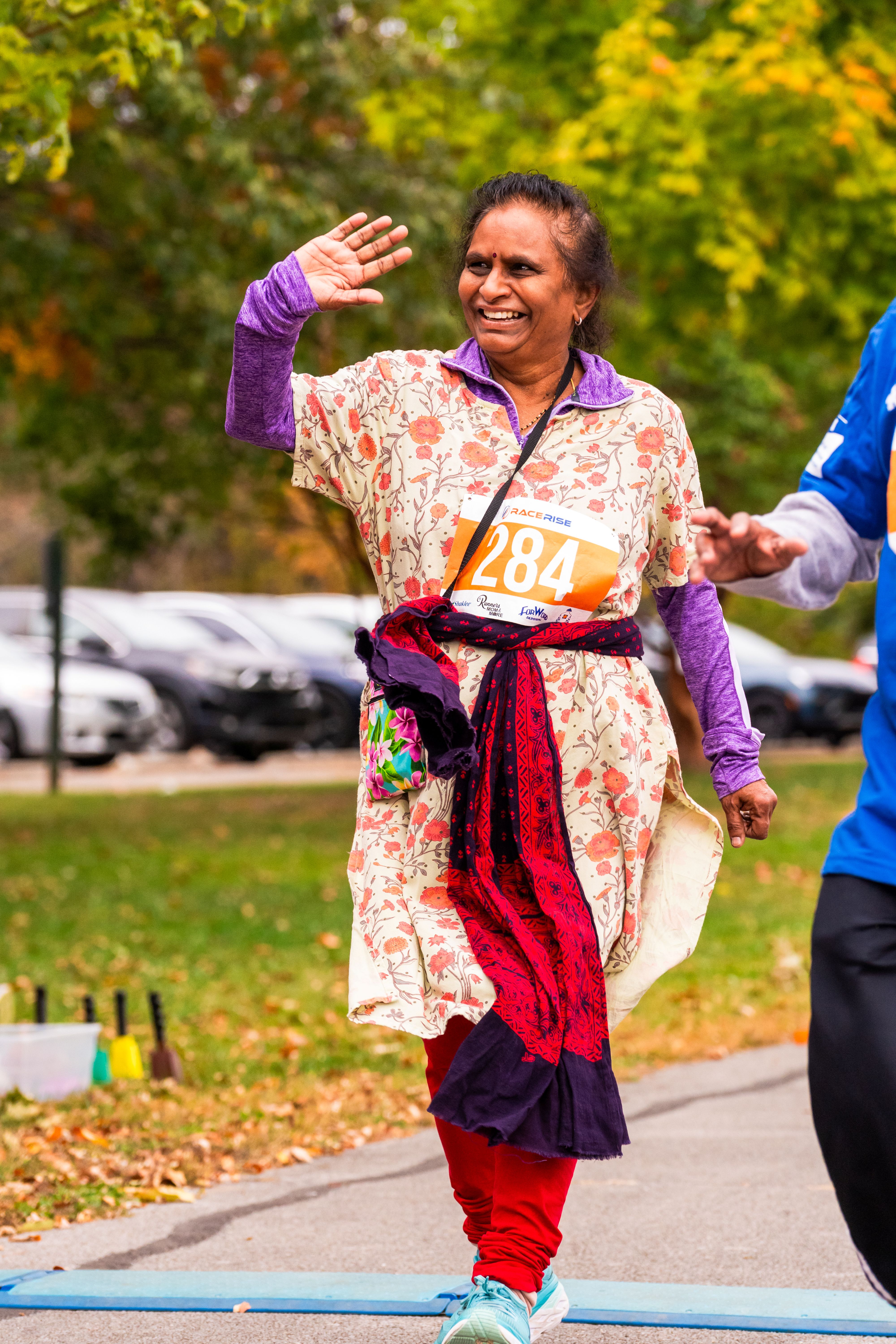 A woman wearing a wrapped garment over a purple, collared shirt waves as she walks across the finish line