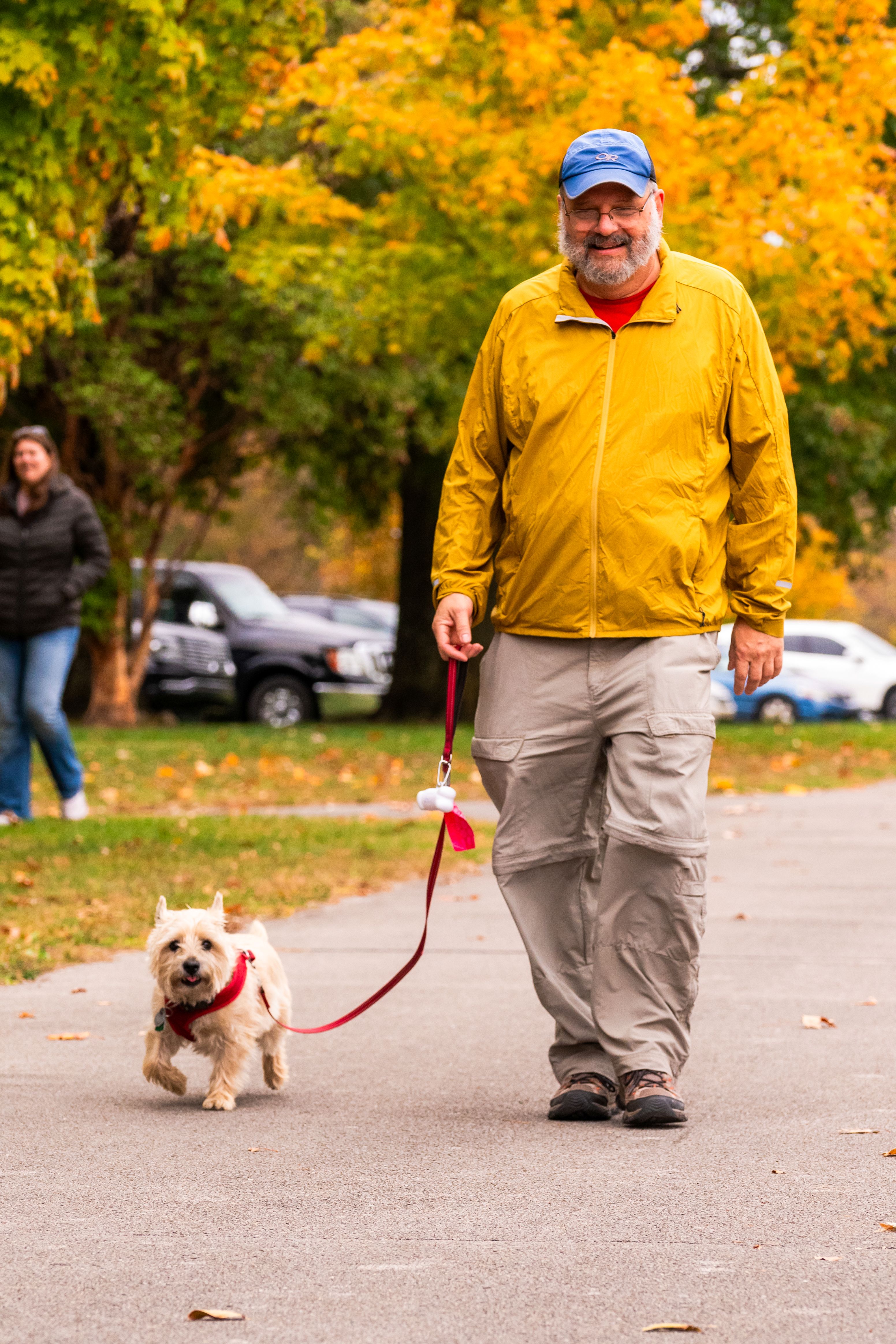 A man in yellow walks a very tiny dog on a red leash. The man is smiling.