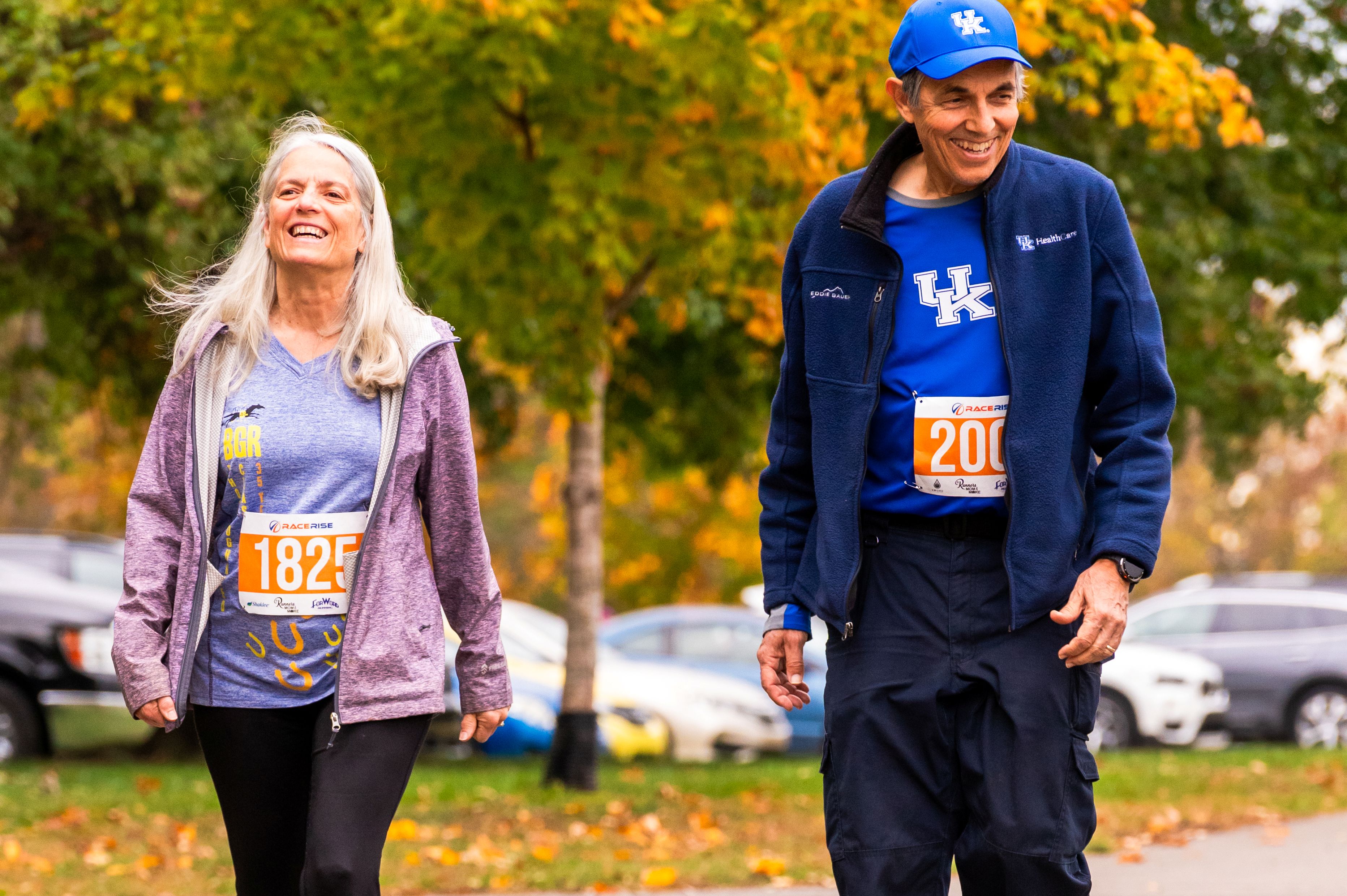 A man and a woman smile while walking. The man wears a University of Kentucky hat and shirt