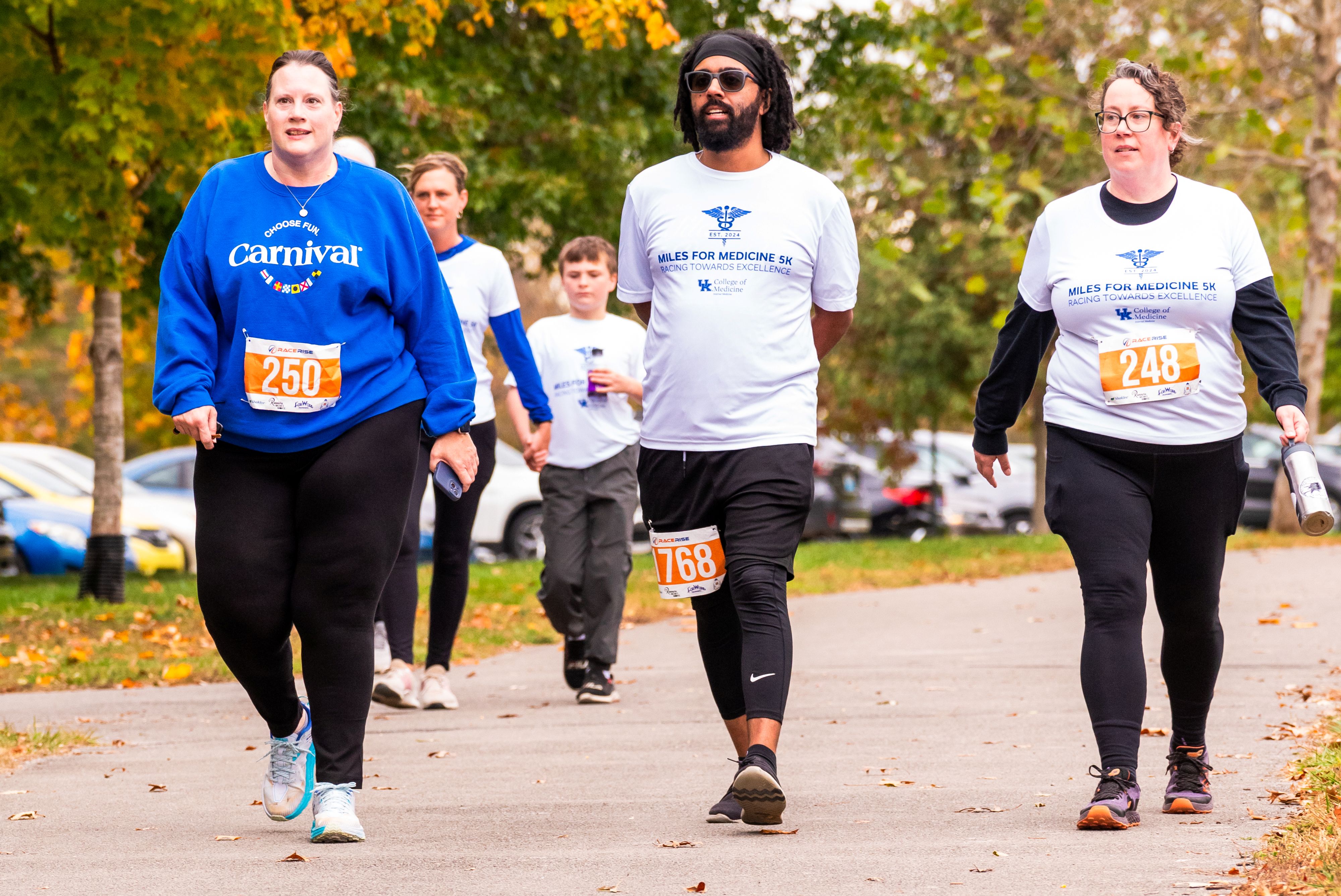 A group of 3 people walk in a line. The man in the middle wears sunglasses.