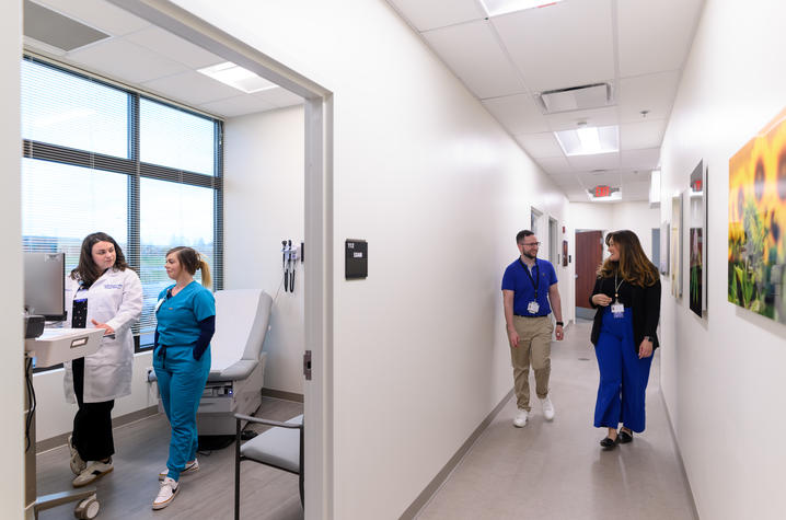 Four people speak at a hospital in two pairs. One pair speaks inside of a hospital room, looking at a computer. The other pair speaks outside in the hallway, smiling.